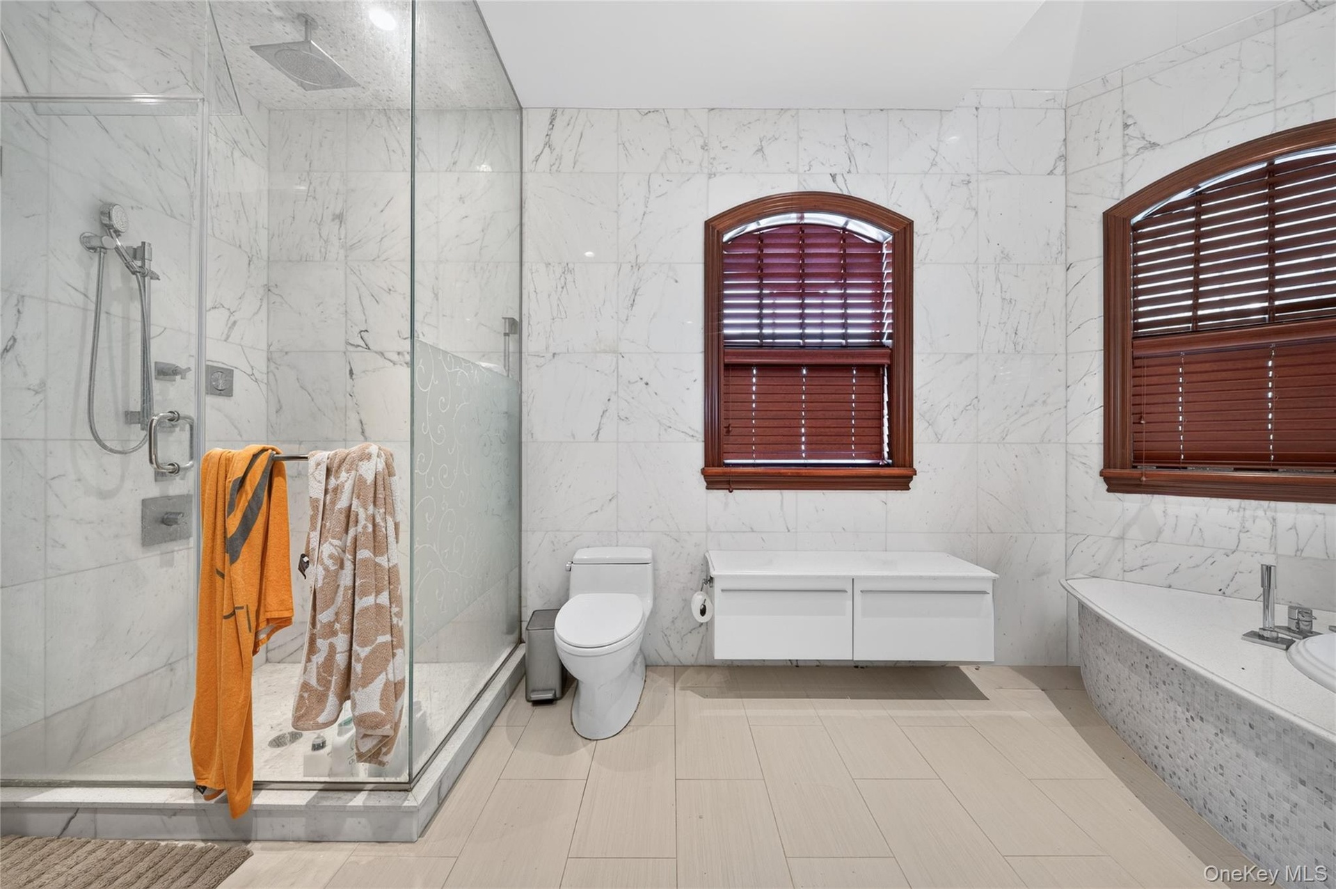 Glass-enclosed shower with multiple showerheads, white tile walls, two arched windows with wood-tone blinds, and a built-in bathtub with a white count