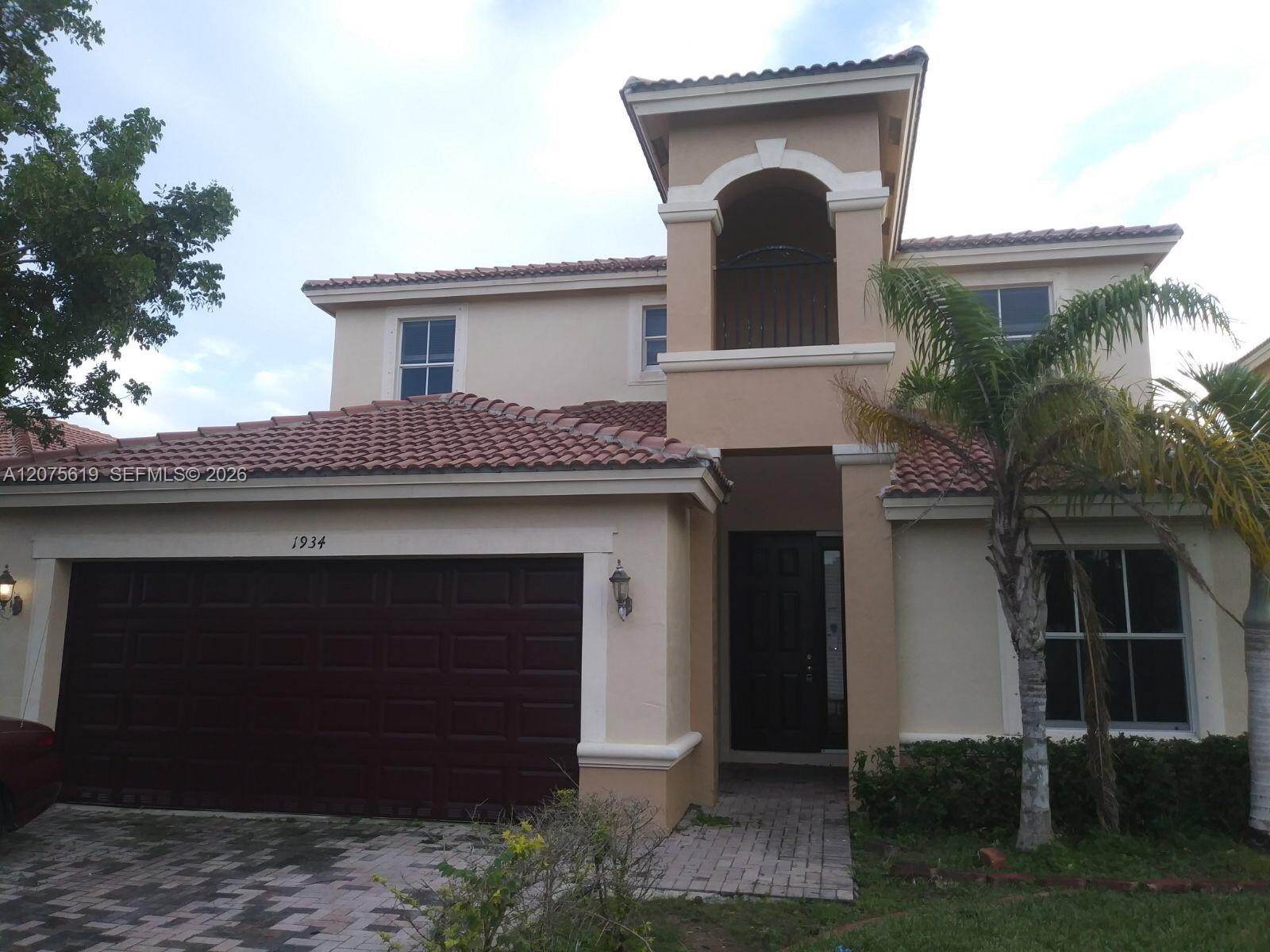 Two-story stucco exterior featuring a tiled roof, paver driveway, attached two-car garage, and established palm landscaping