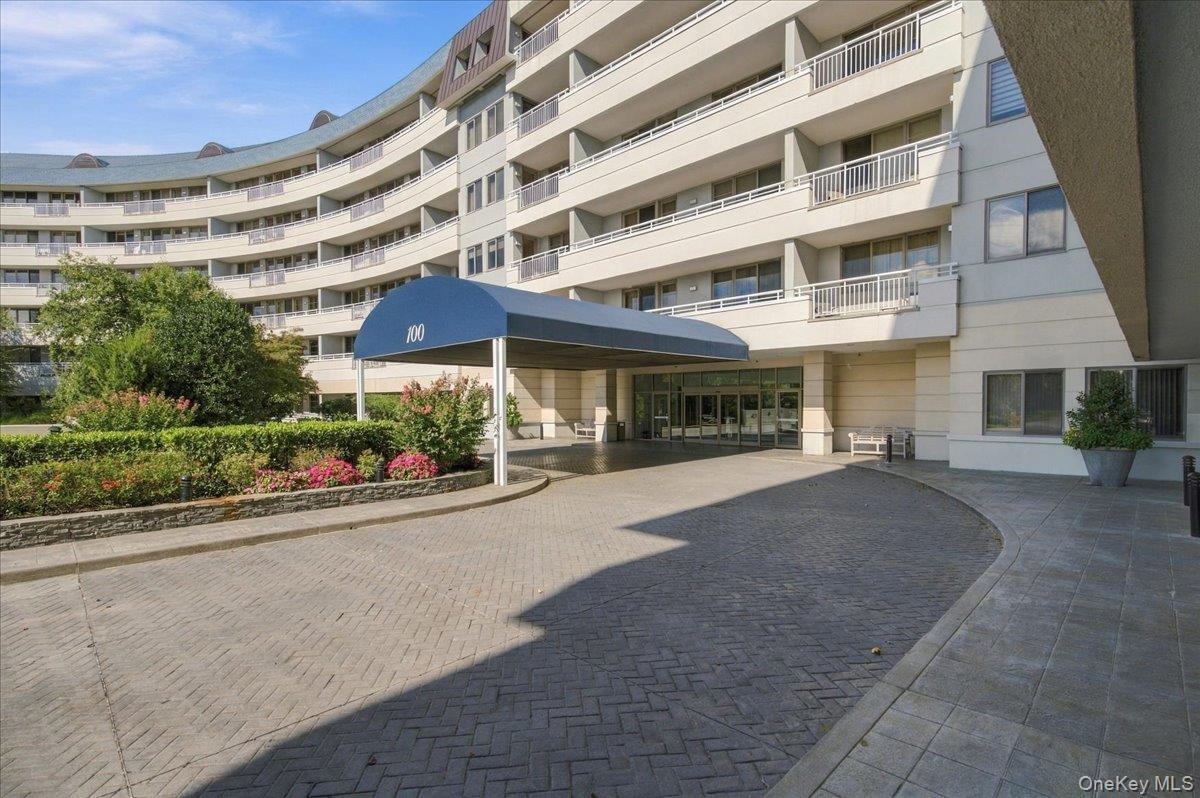 Curved multi-story building exterior with individual balconies, covered entryway with a blue canopy, and a paver driveway