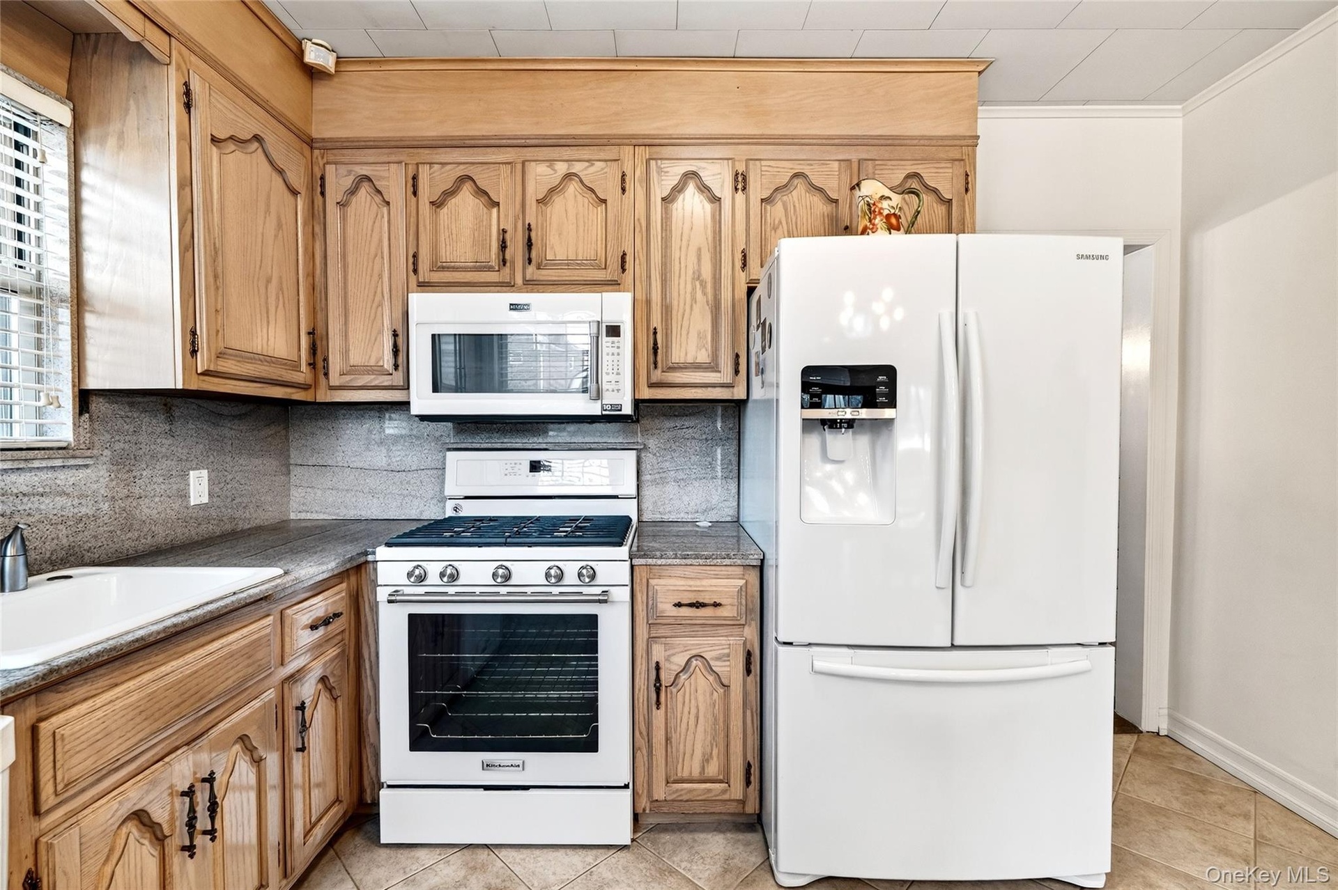 Kitchen with white appliances, backsplash, light tile patterned flooring, dark stone counters, and brown cabinets