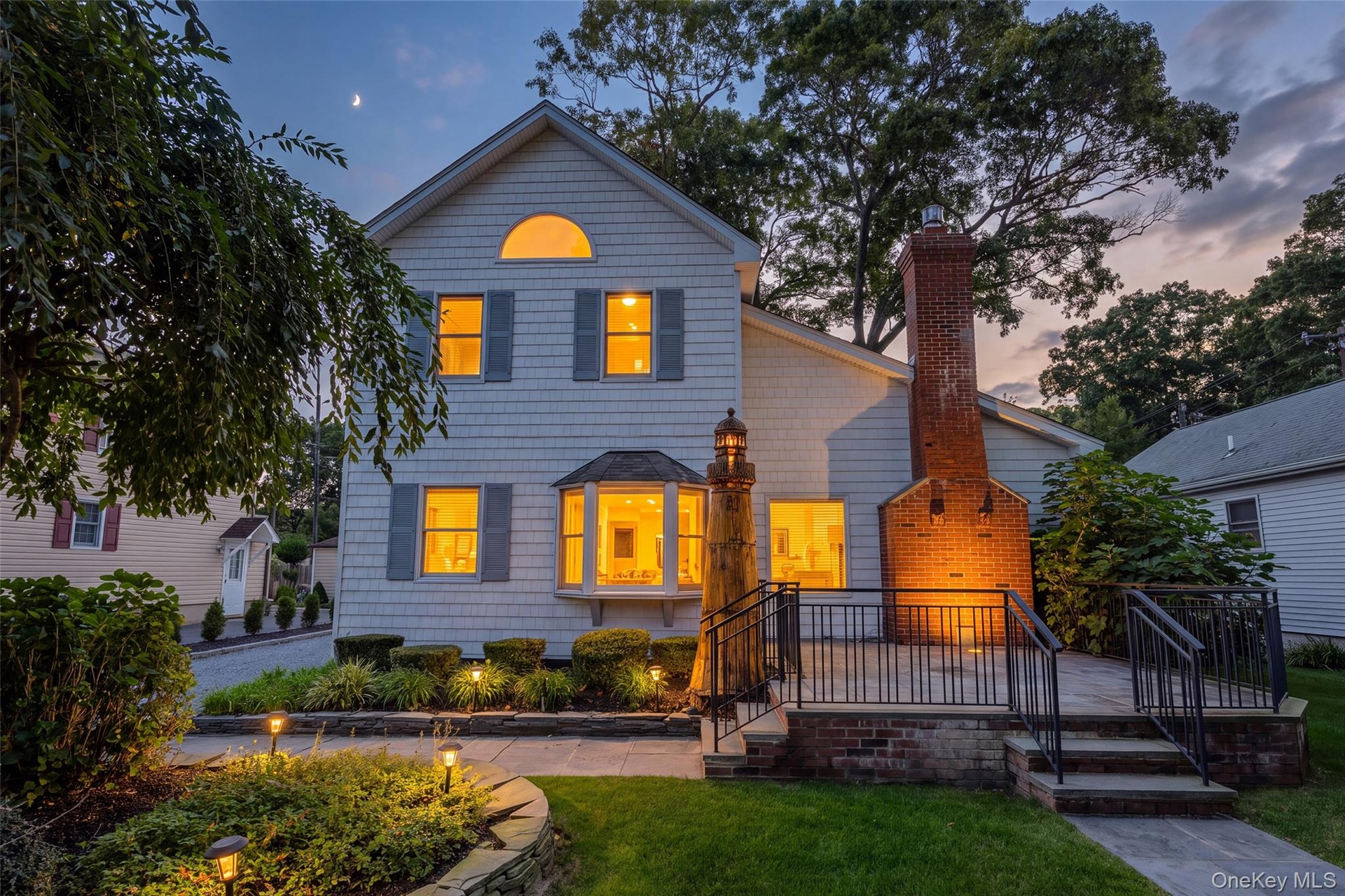 White shingle siding with gray shutters, bay window, prominent brick chimney, multi-level patio with black railings, and stone-edged landscaping with 