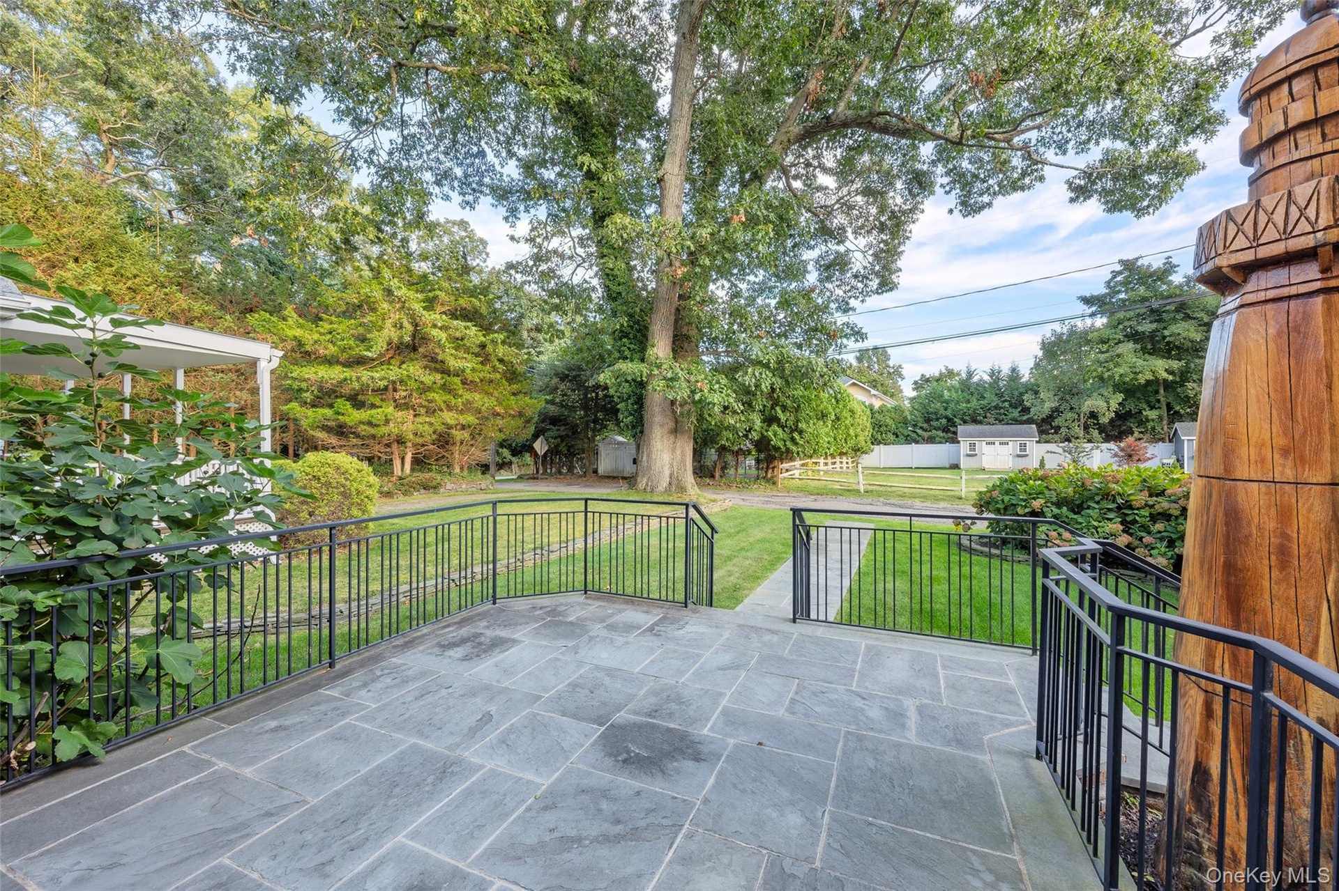 Stone patio with black iron railings, a manicured lawn, and mature trees