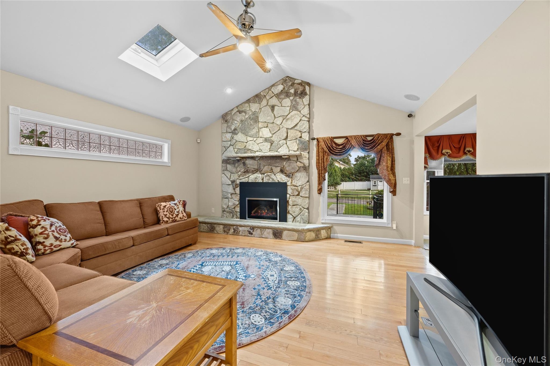 Vaulted ceiling with skylight, stone-clad fireplace, wood-finish flooring, and a ceiling fan