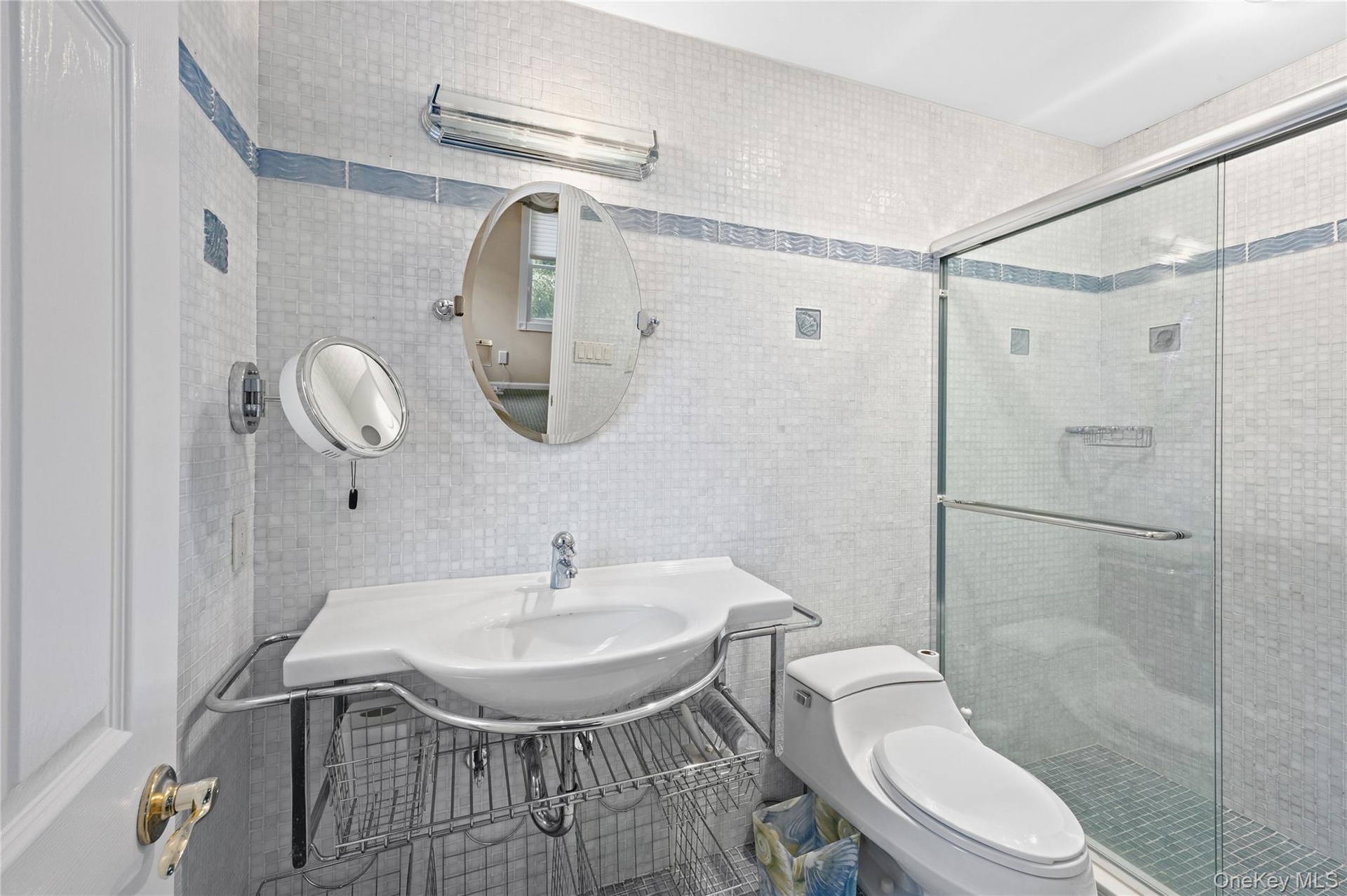 Shower with glass enclosure, white square tile walls with blue accent banding, white pedestal sink with chrome-finish basin, and oval wall mirror