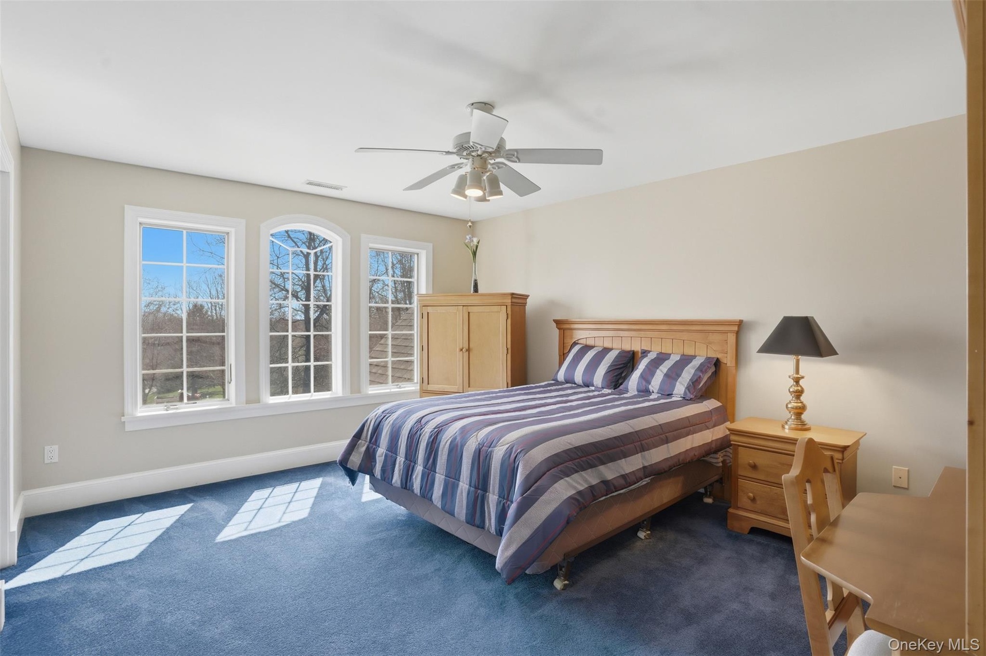 This bright bedroom features a ceiling fan, plush blue carpet, and a trio of windows offering views of the outdoors