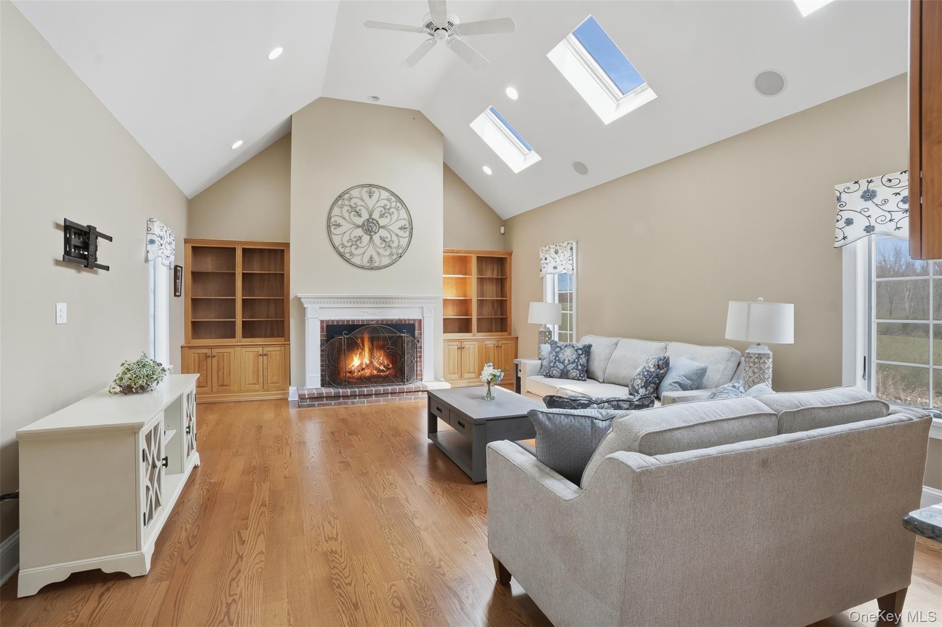 Spacious living area featuring vaulted ceilings, hardwood floors, a brick fireplace with a white mantel, built-in wooden shelving, and skylights provi