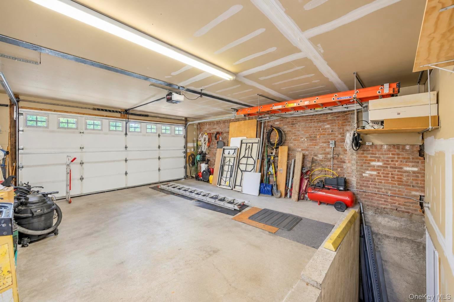 2 car garage, Concrete flooring, exposed brick wall, overhead lighting, segmented garage door with windows, and an overhead storage rack