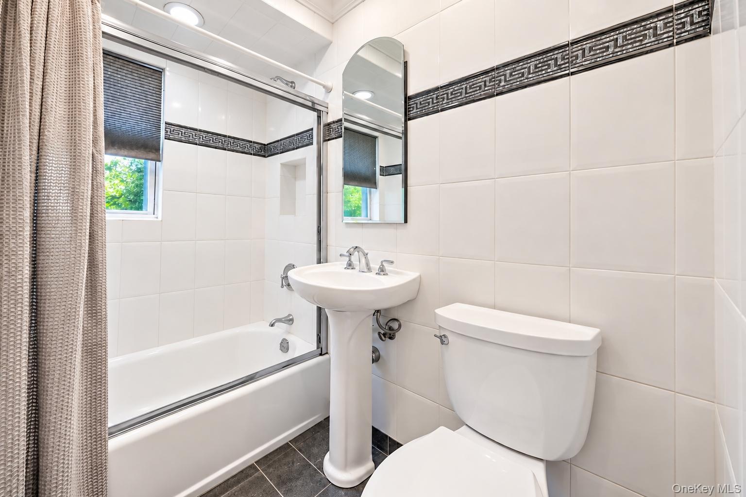 White tile walls featuring a decorative border, pedestal sink, and a built-in bathtub with a glass enclosure