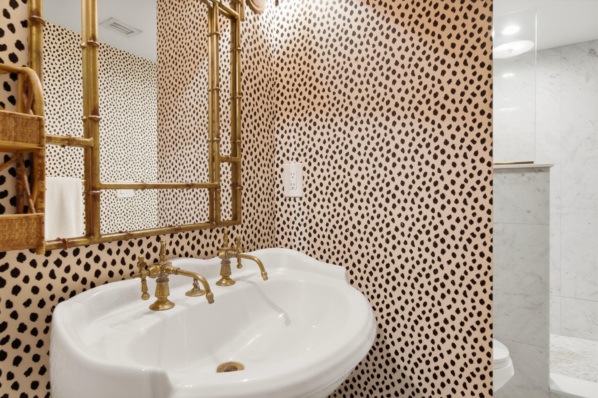 Pedestal sink with brass hardware, framed mirror, patterned wallpaper, and a glass-enclosed shower with tiled walls