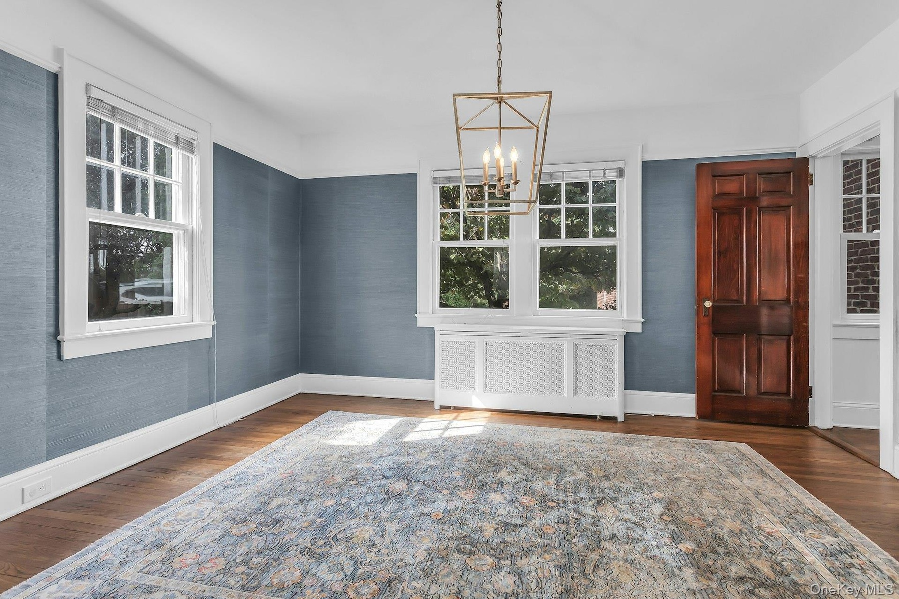 Hardwood flooring with a matte finish, blue textured wall covering, a dark wood panel door, white window casings, and a geometric chandelier