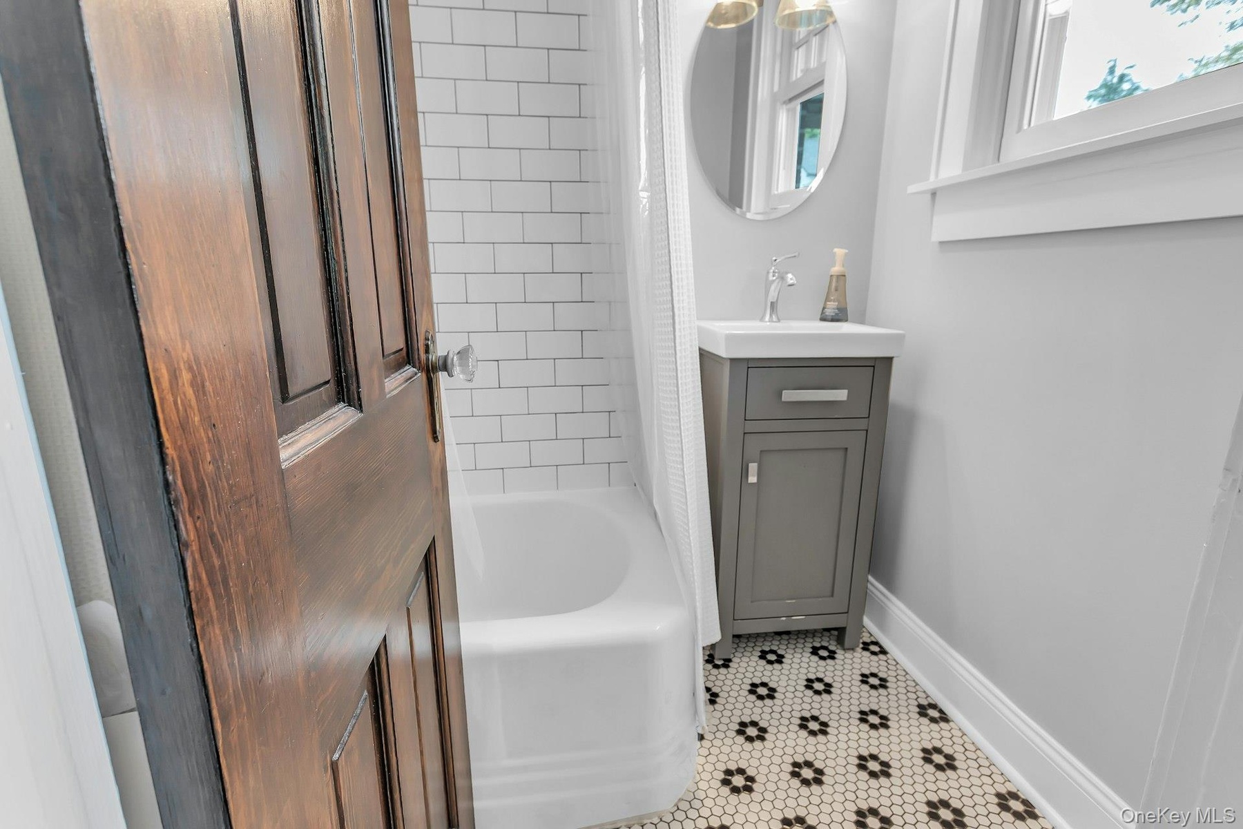 Subway tile bath surround, white tub, gray vanity with integrated sink, patterned mosaic floor tile, and an oval mirror
