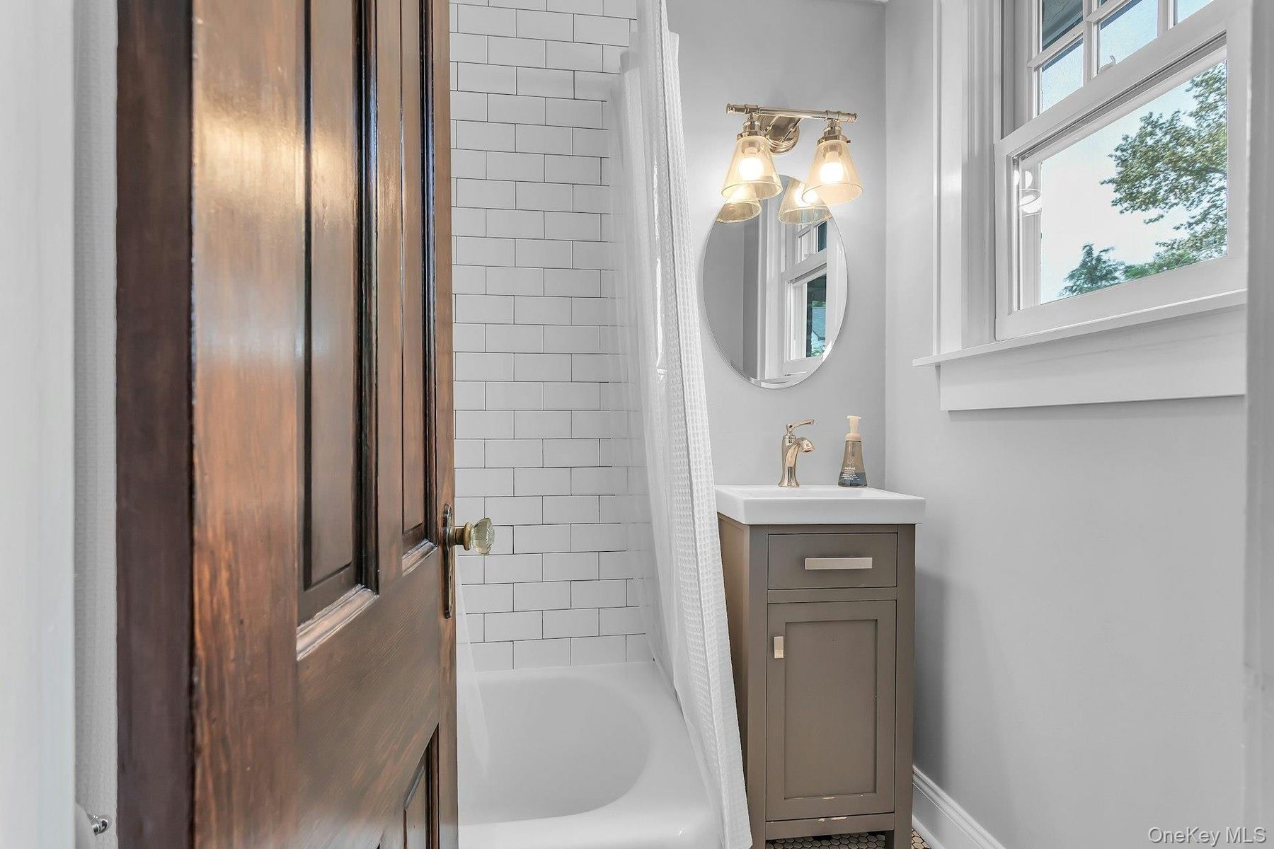 White subway tile shower surround, single window with white trim, gray vanity with integrated sink, gold-toned faucet, and oval wall mirror