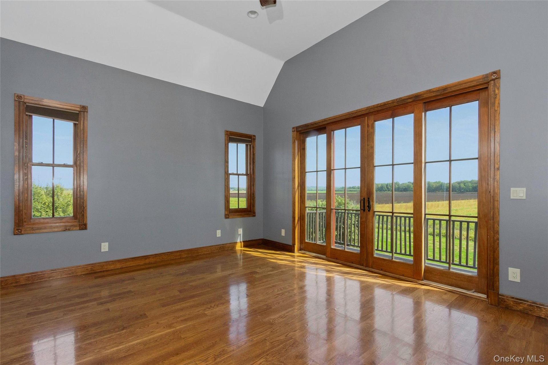 Hardwood flooring, vaulted ceiling, two windows with wood trim, and a four-panel sliding glass door with wood trim opening to a balcony with black rai