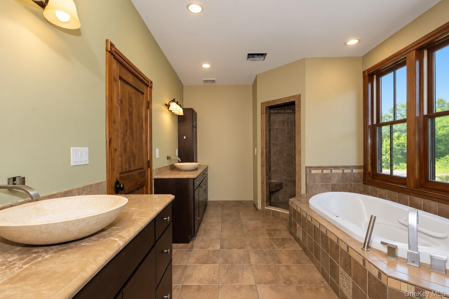 Dual vanity with vessel sinks and stone countertops, a built-in soaking tub, a tiled walk-in shower, and a large window with wood framing
