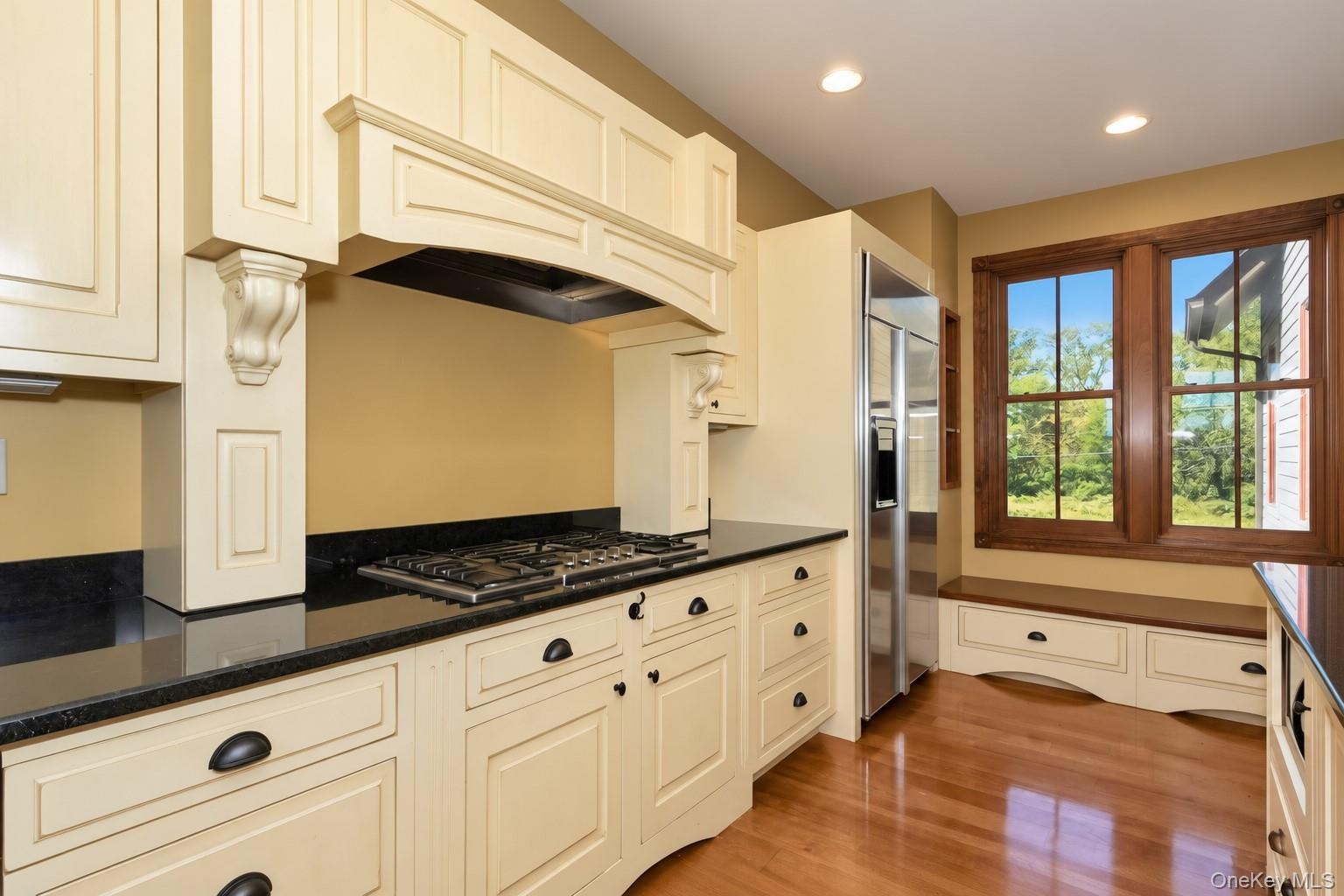 Cream-painted cabinetry, dark countertops, built-in window seating, stainless steel refrigerator, and wood-finish flooring