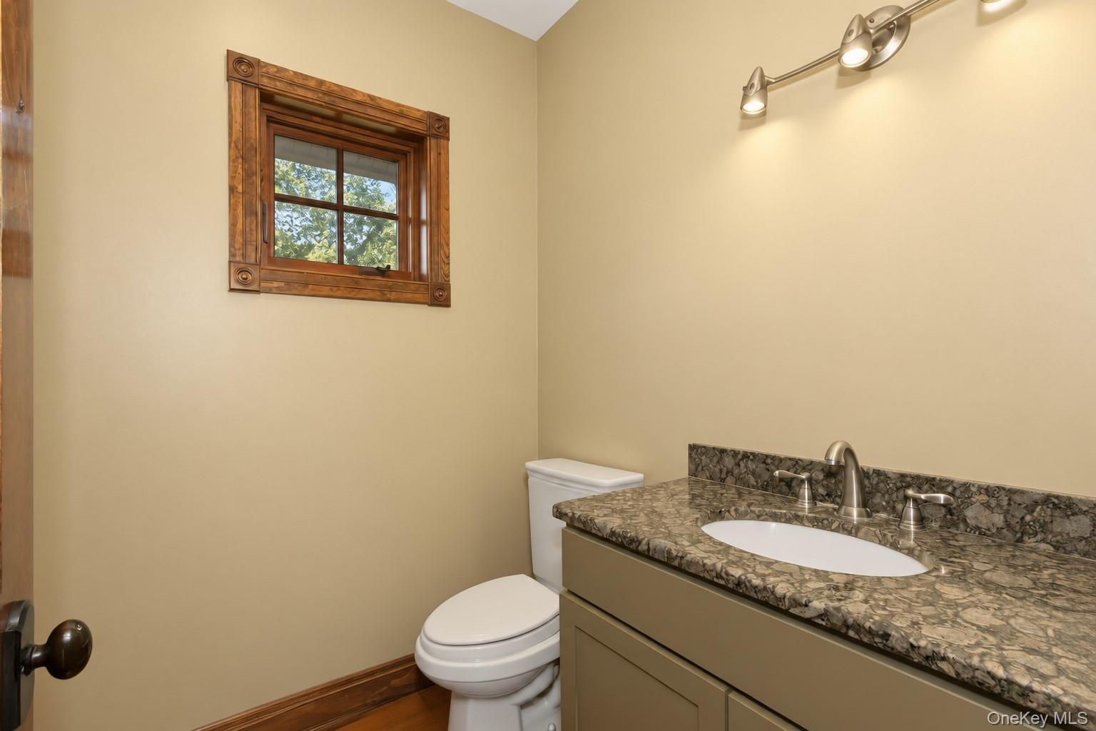 Vanity with a stone countertop and undermount sink, brushed nickel faucet, wall-mounted light fixture, and a window with wood trim