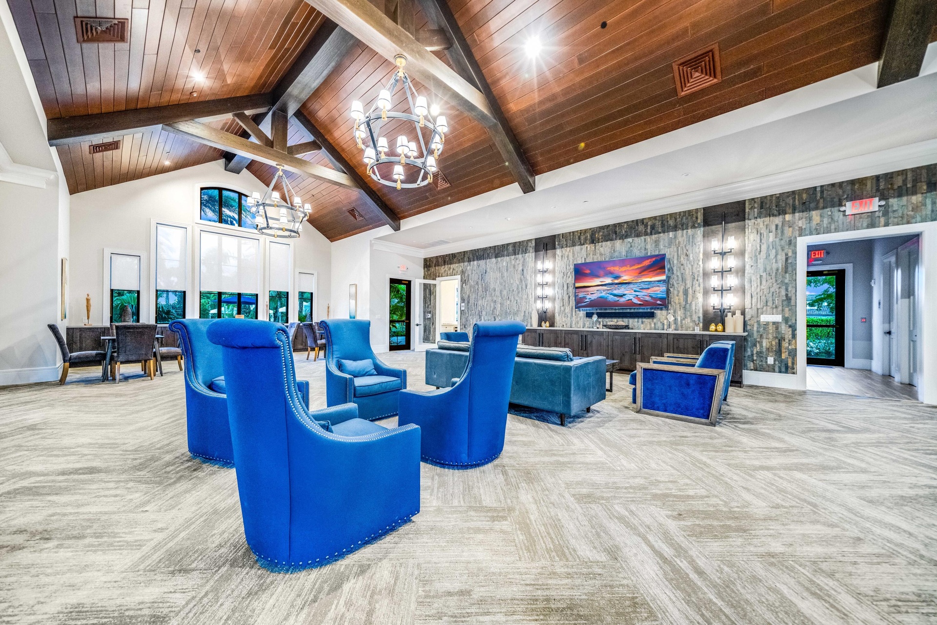 Vaulted wood-paneled ceiling with exposed beams, dual chandelier lighting, floor-to-ceiling windows, stone accent wall, and patterned carpet flooring