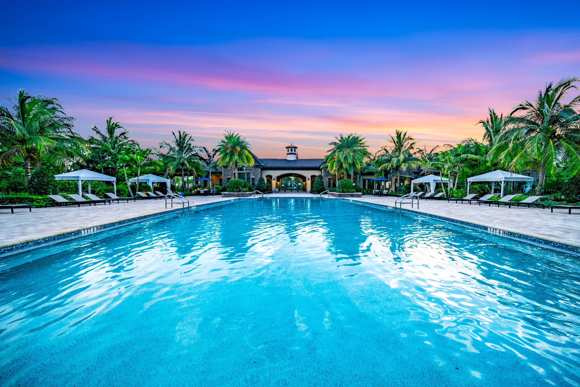 Expansive outdoor pool with a surrounding stone deck, tropical landscaping, and a distant clubhouse structure