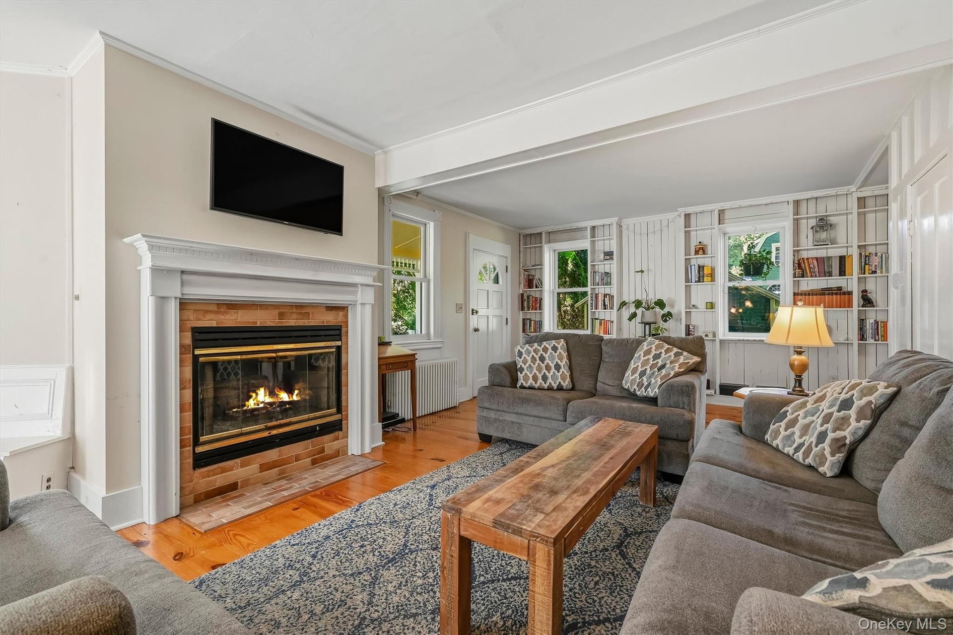 Fireplace with a white mantle and brick surround, hardwood flooring, built-in shelving, and white trim work