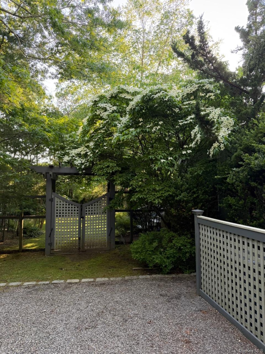Gravel drive with a backyard entrance with lattice fence and gate, featuring mature trees and flowering shrubbery