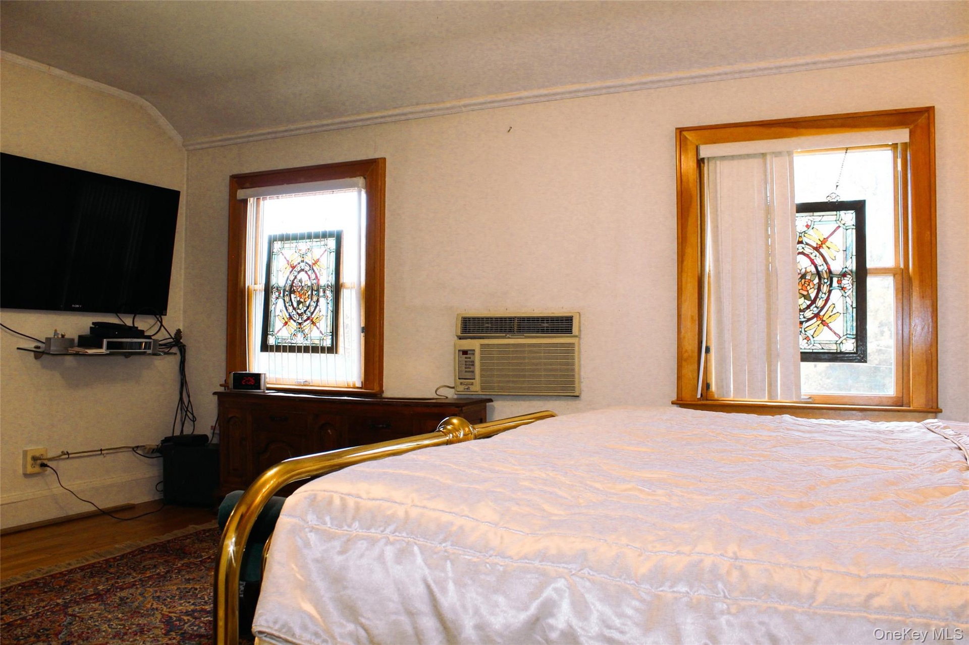 Large bedroom 1 with wood-frame windows with stained glass decorative inserts, white textured walls, and a wall-mounted air conditioning unit