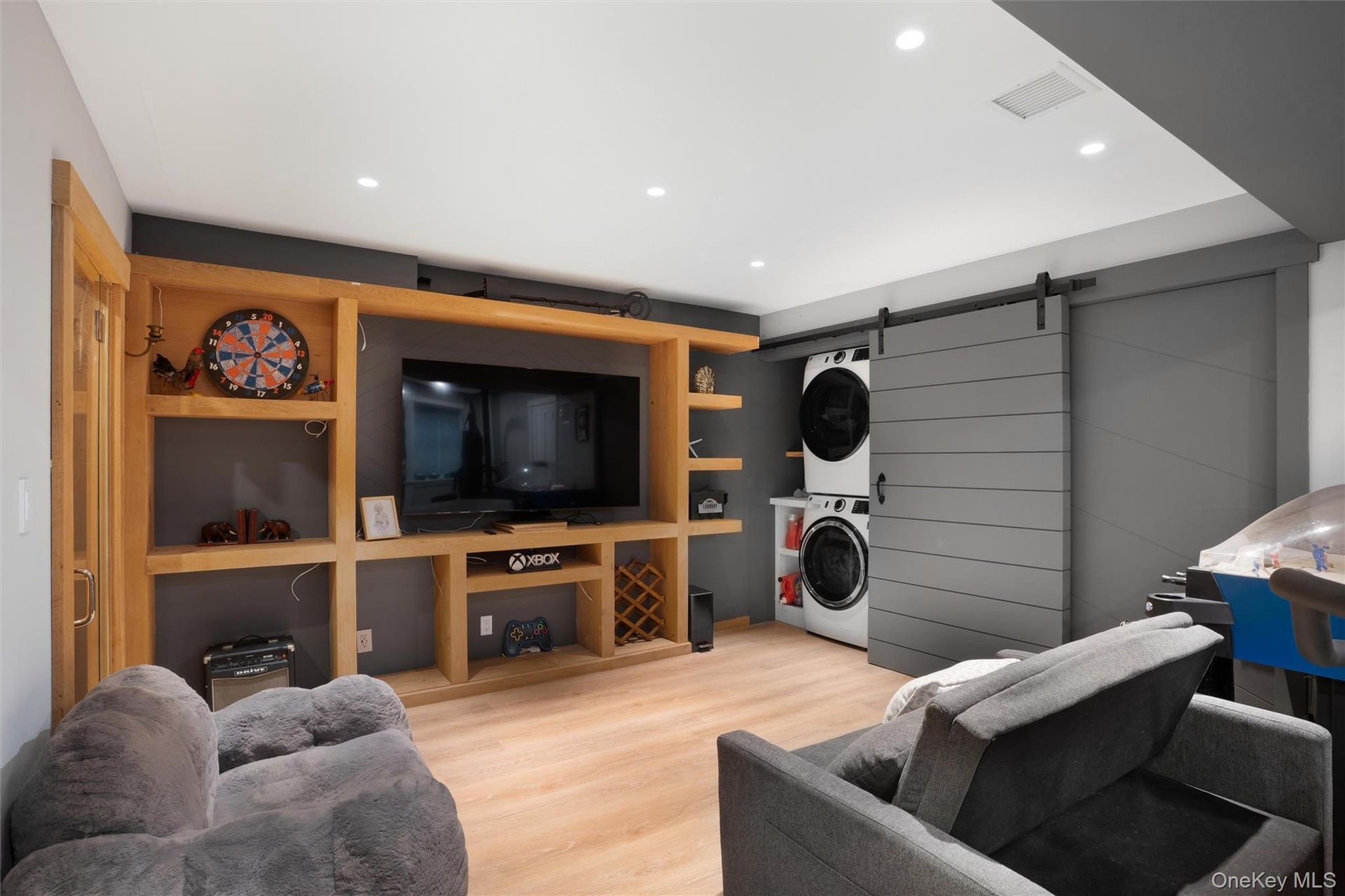 Built-in entertainment center with open shelving, light wood-finish flooring, recessed lighting, and a barn door concealing stacked laundry appliances