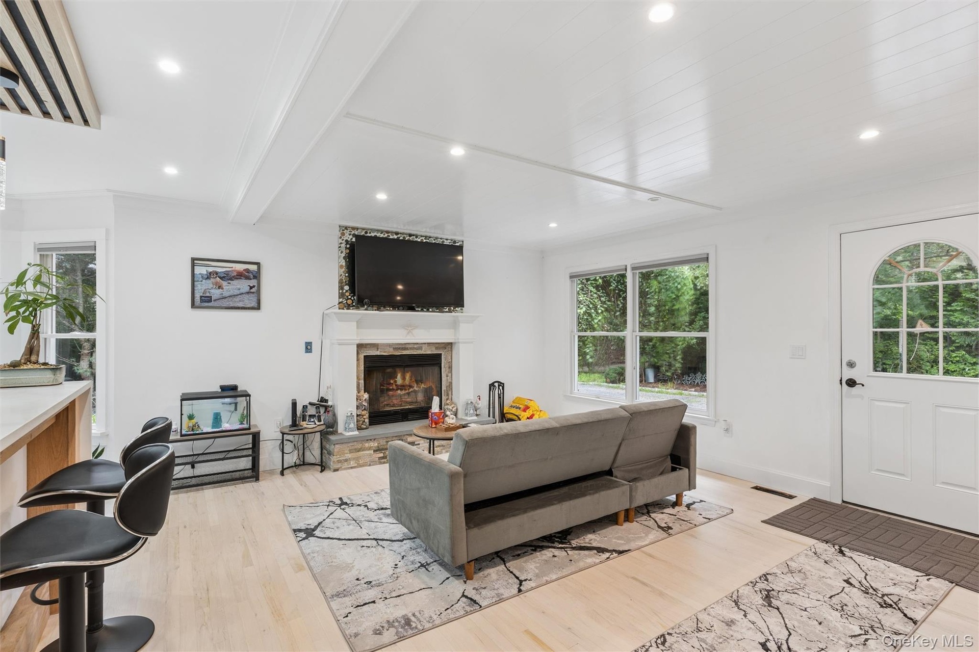 Vaulted ceiling with exposed beams, recessed lighting, wood-finish flooring, stone fireplace, and a white paneled entry door with an arched window
