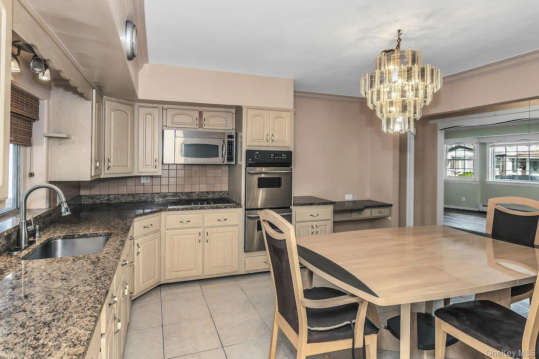 Kitchen featuring light-toned cabinetry, granite-finish countertops, stainless steel appliances, a tile backsplash, and a contemporary chandelier