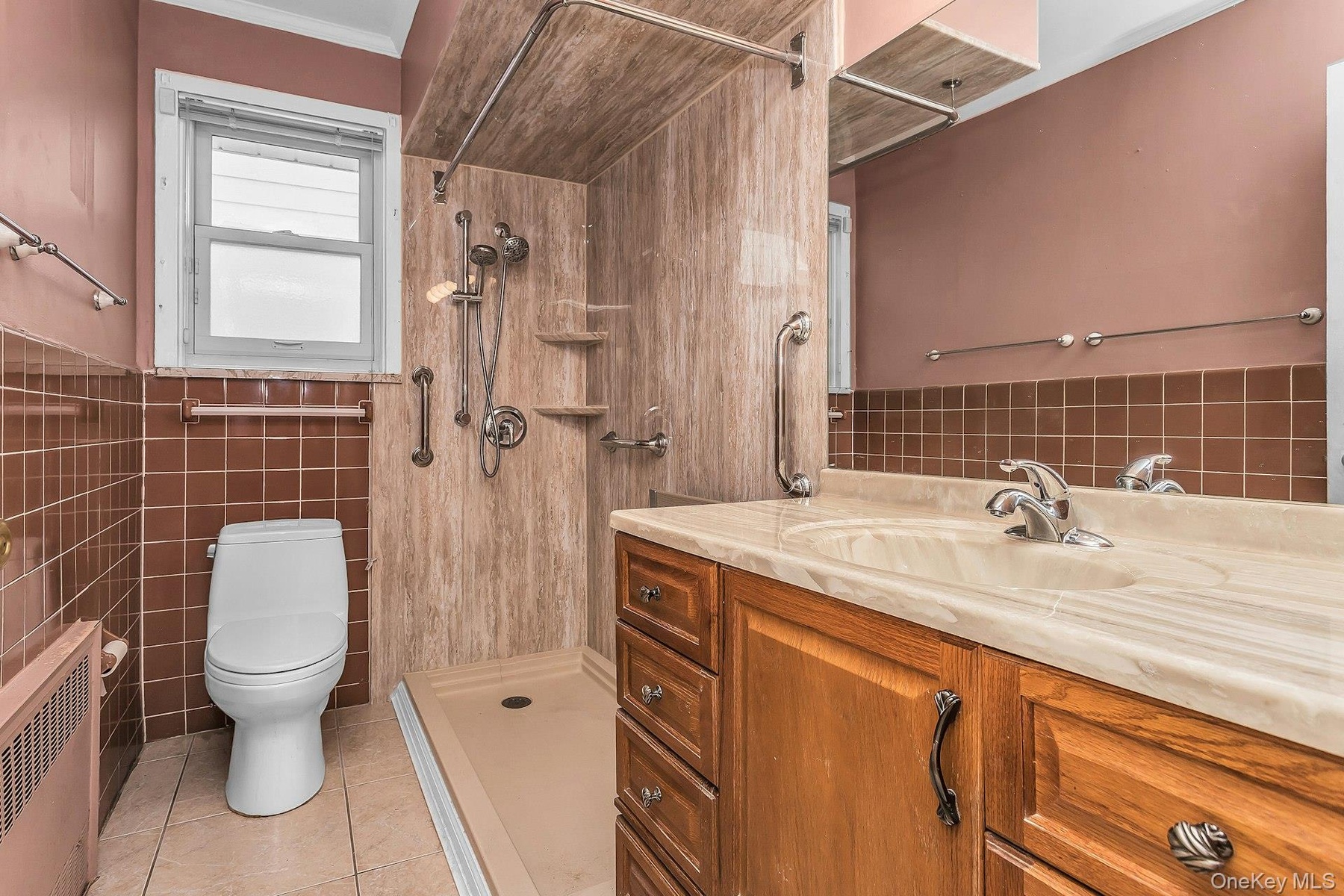 Bathroom featuring a wood-finish vanity with an integrated sink, a large mirror, a tiled shower with grab bars, and a window