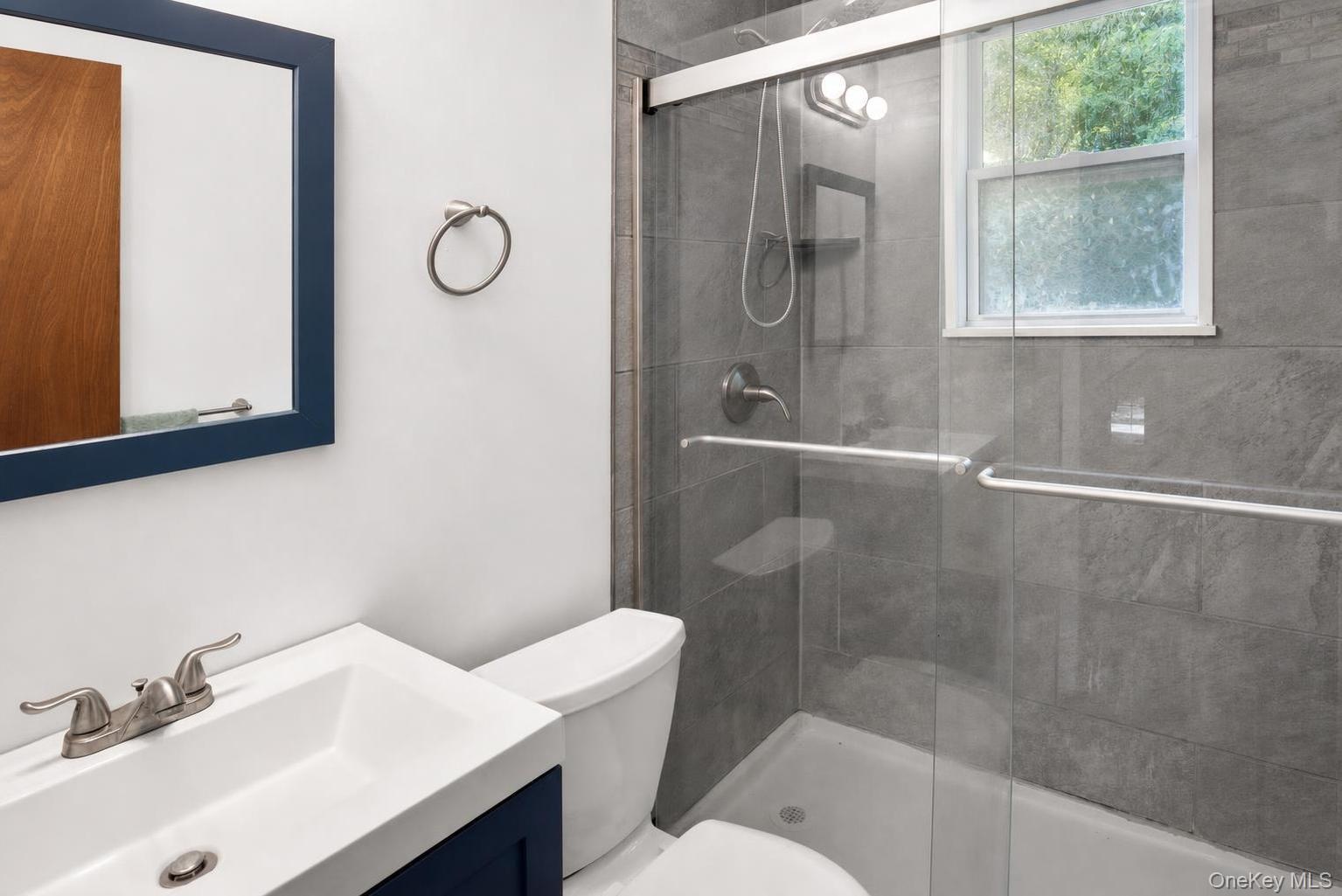 Bathroom featuring a vanity with an integrated sink, a toilet, a shower with a sliding glass door, grey tile surround, and a window