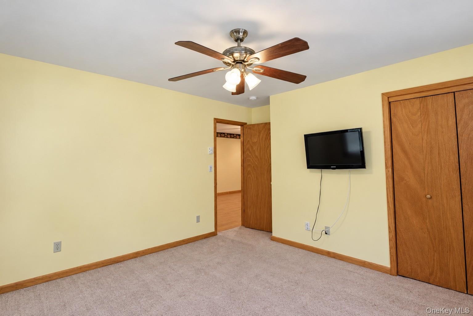 Ceiling fan with integrated lighting, wood-finish bi-fold closet doors, wall-mounted television, and neutral carpeting