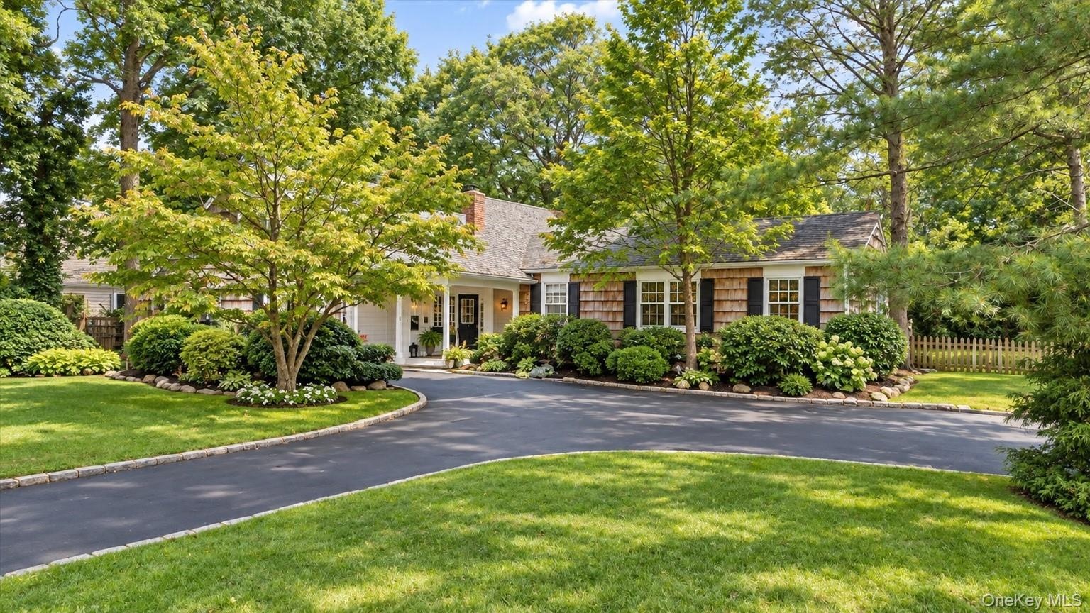 Asphalt driveway, manicured lawn, shaker siding, mature trees, and a covered entry