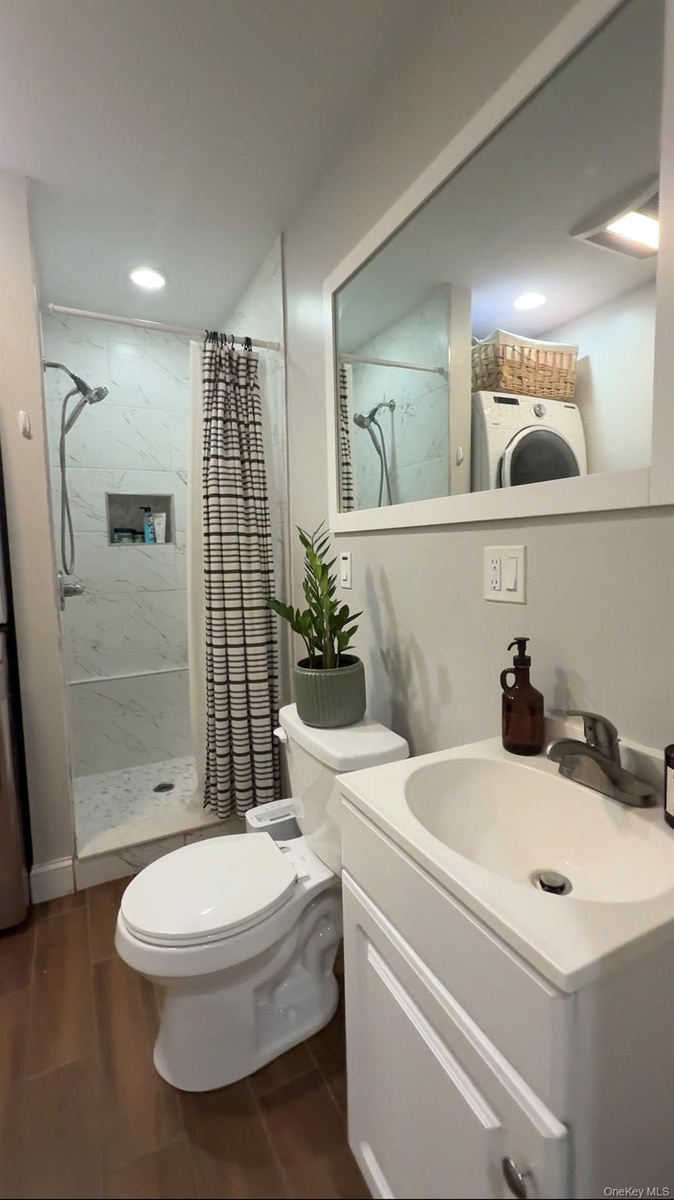 Cottage full bathroom with wood-finish flooring, stacked laundry appliances, walk-in shower with glass enclosure, recessed lighting, and a vanity with
