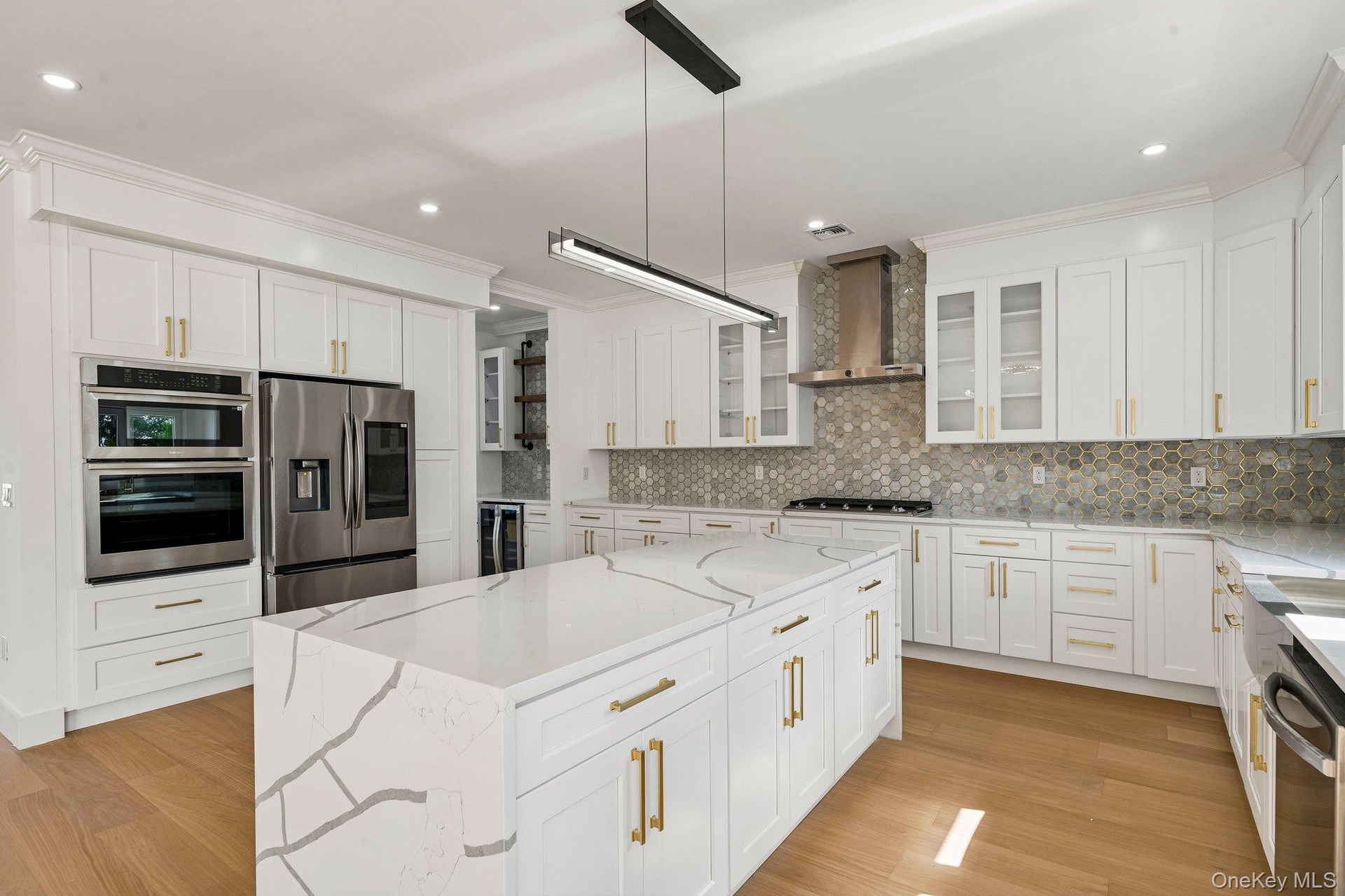 Gourmet kitchen featuring a waterfall island, stainless steel appliances, honeycomb tile backsplash, white cabinetry with gold-tone hardware, and wood