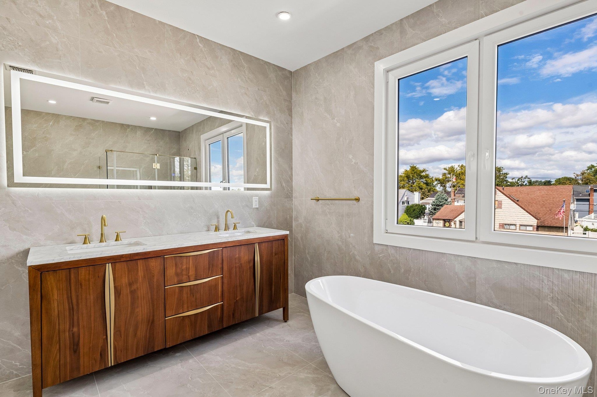 Freestanding soaking tub, dual-pane windows, wood-finish double vanity with stone countertop, and an illuminated mirror