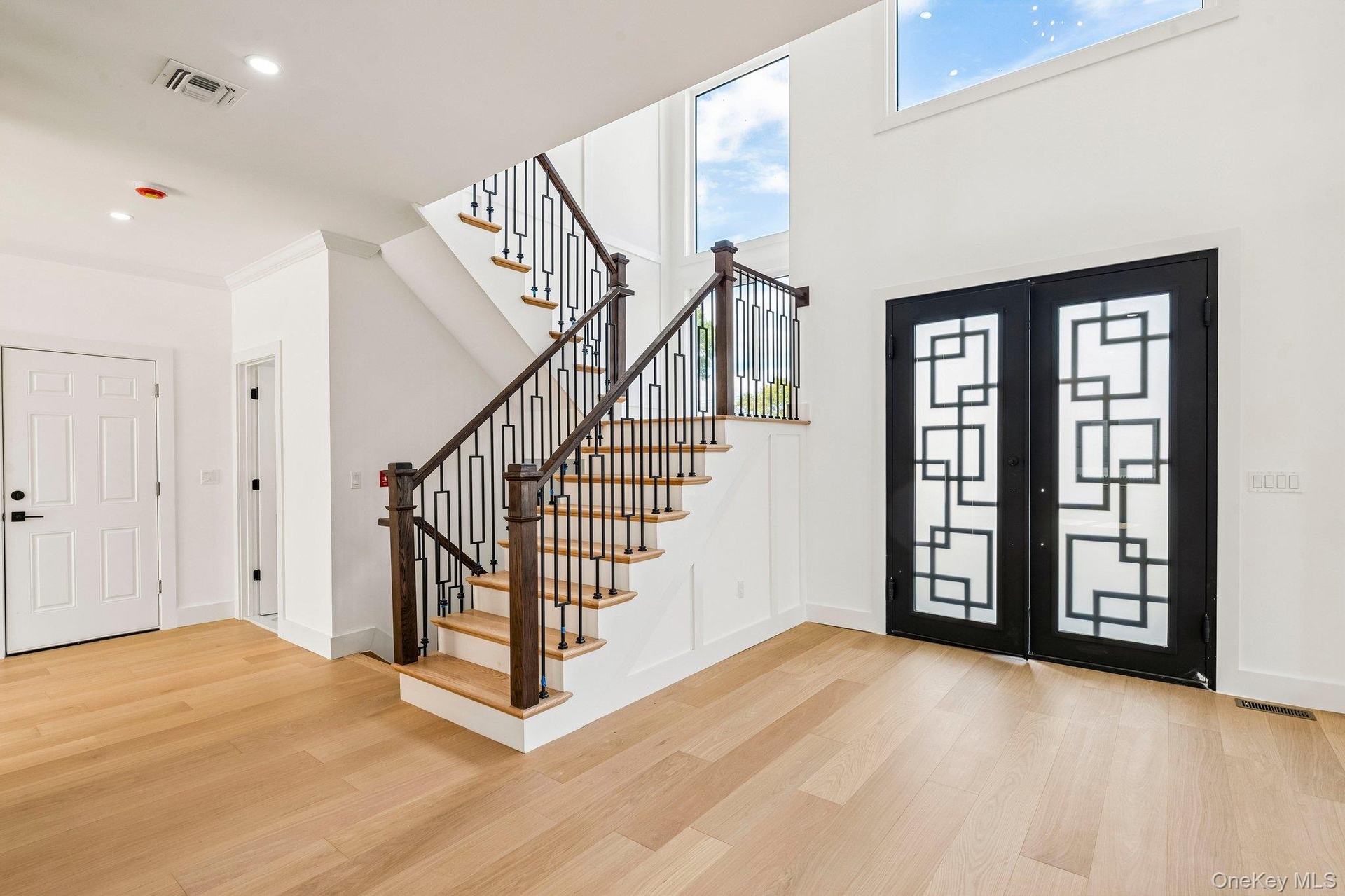 Two-story entryway with light wood-finish flooring, a contemporary black metal and wood staircase, and double entry doors with geometric glass inserts