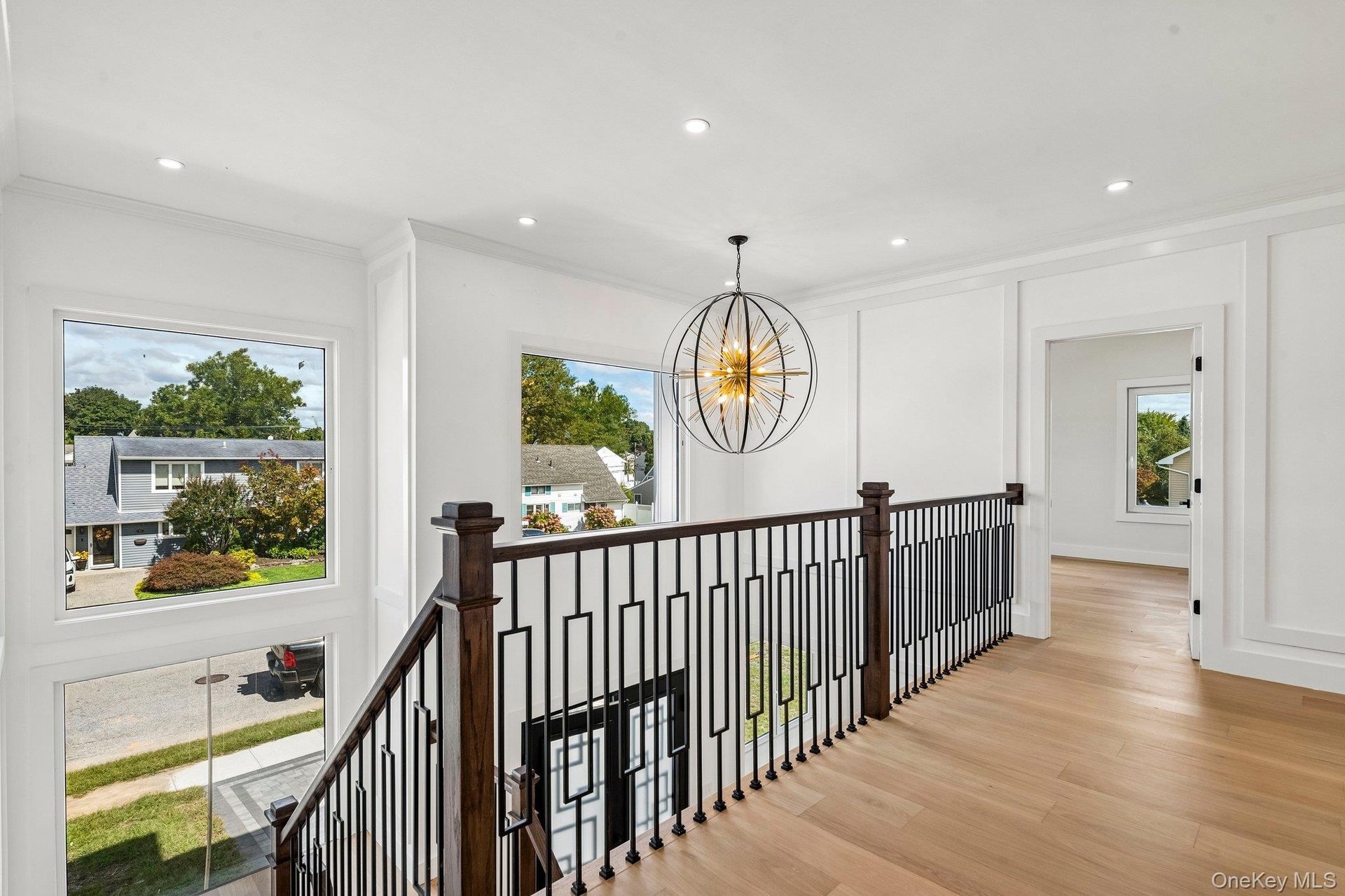 Hardwood flooring, custom iron balusters, recessed lighting, white wall paneling, and an oversized chandelier