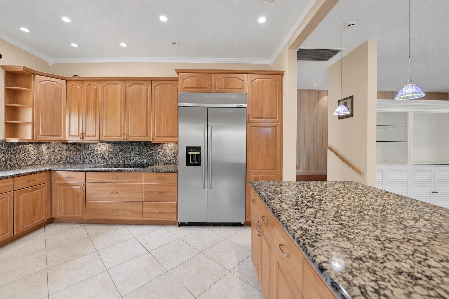 Kitchen featuring extensive wood cabinetry, stainless steel refrigerator, patterned stone countertops, recessed lighting, and tile flooring. Wall colo