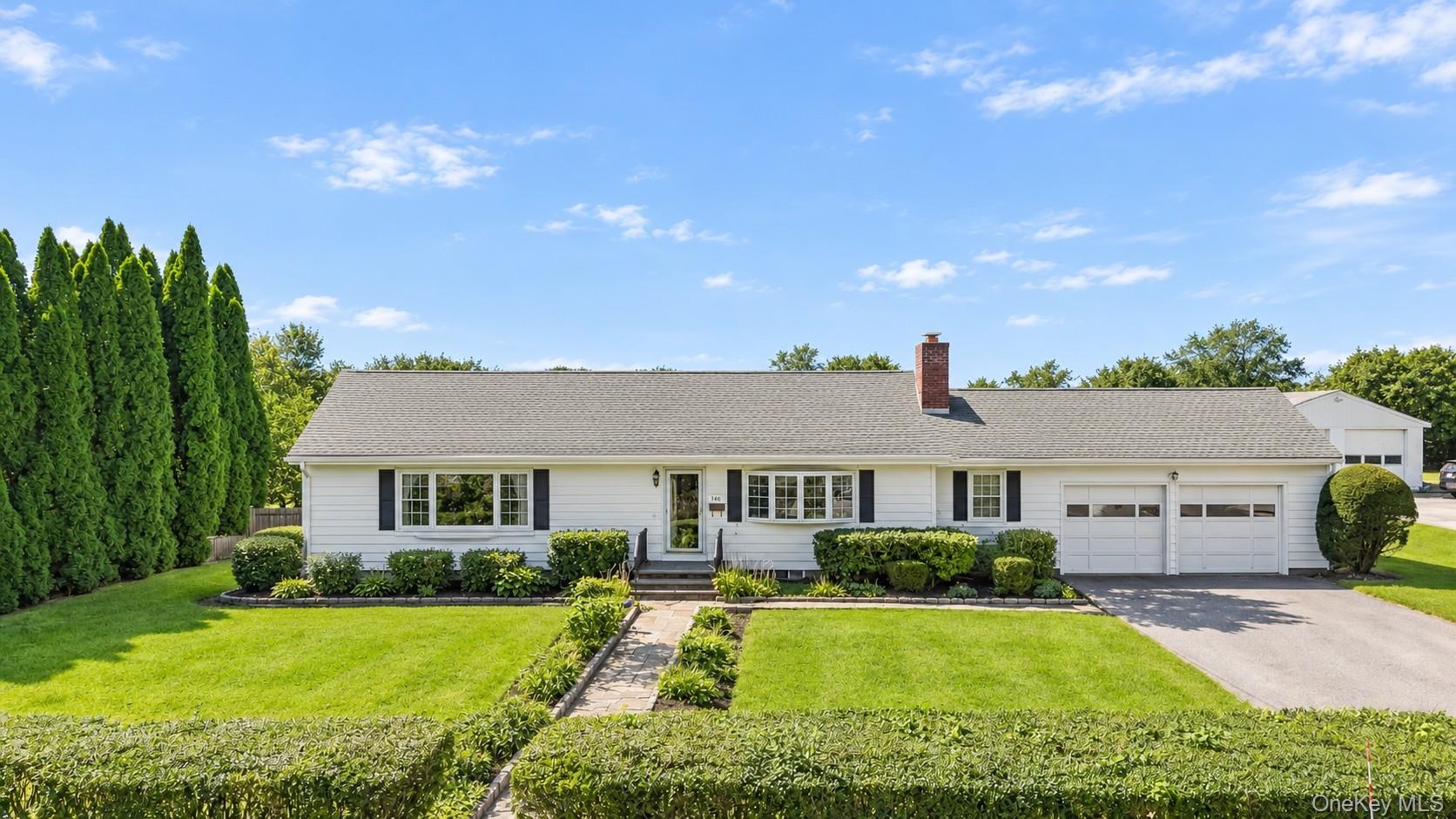 White ranch-style residence featuring a two-car garage, brick chimney, established front landscaping, and a paved driveway