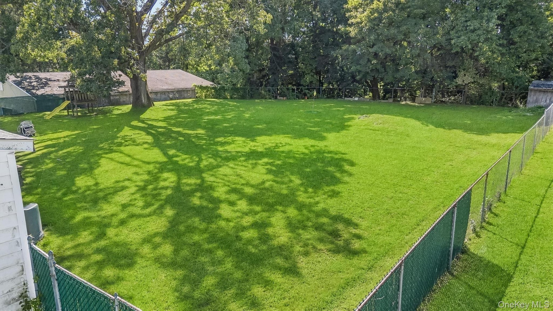 Expansive grassy yard featuring a chain-link fence, mature trees, and a shaded area