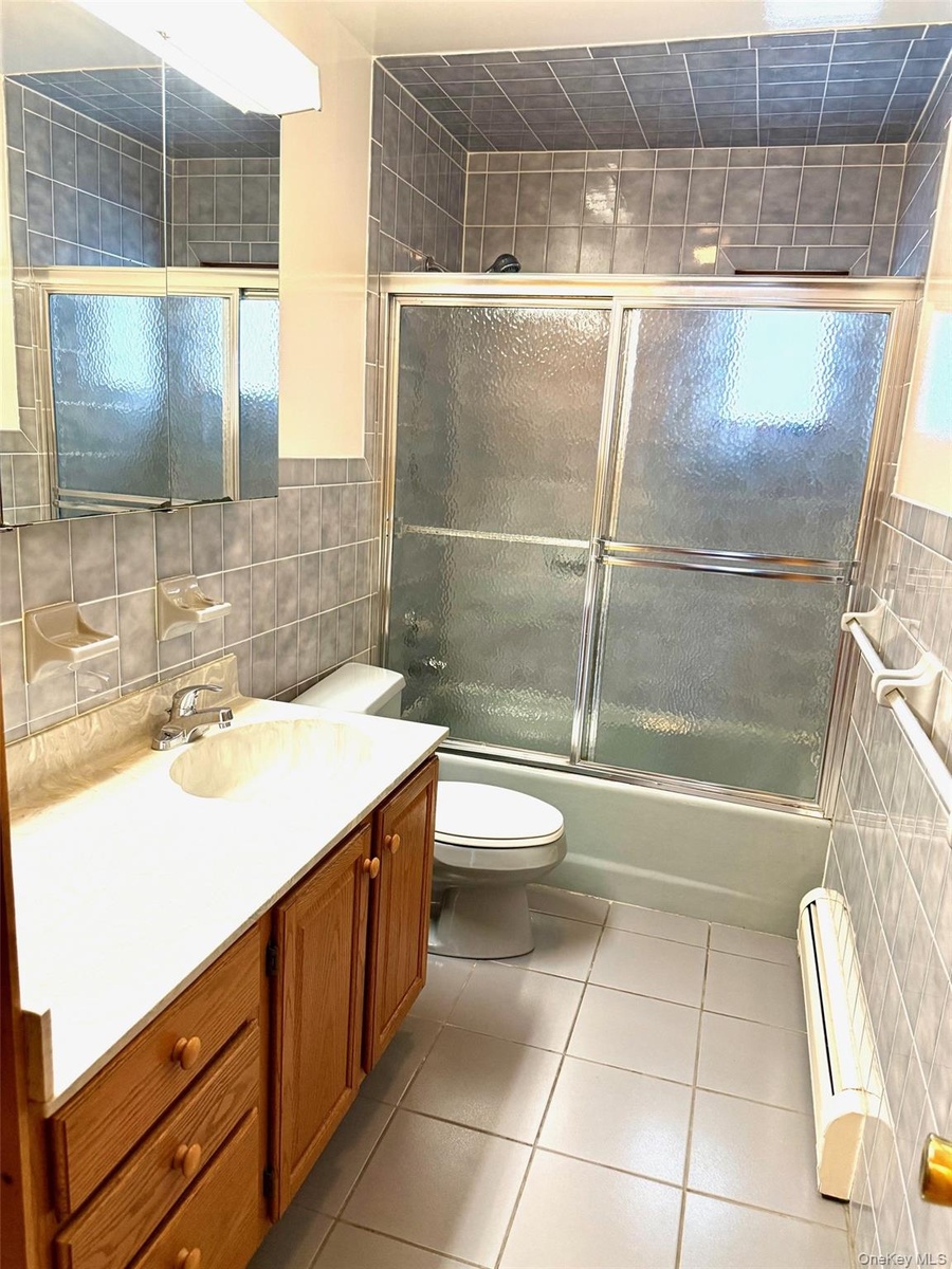 Hall Bathroom with tile walls, shower / bath combination with glass door, vanity, light tile patterned floors, and a baseboard radiator