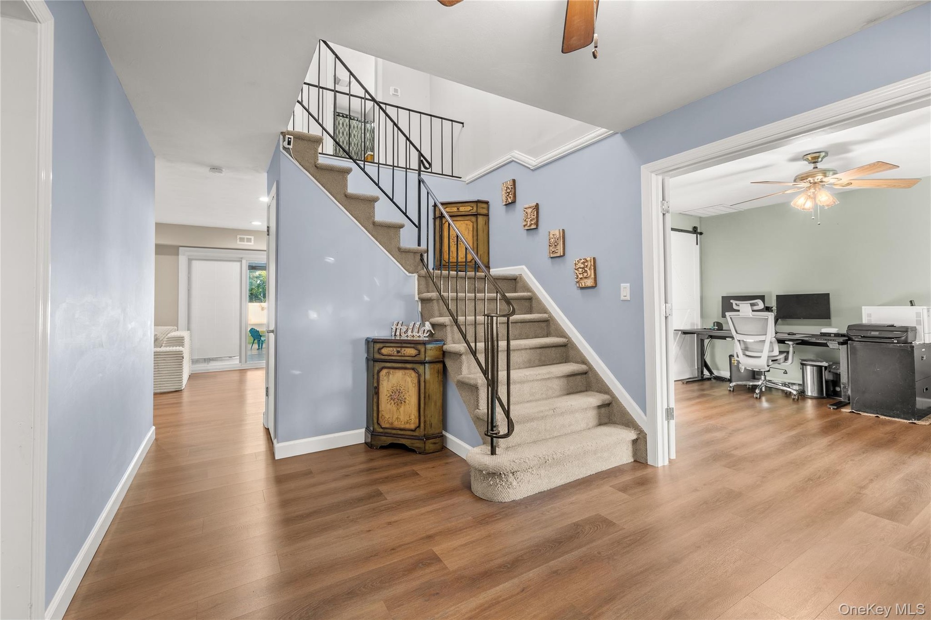 Wood-finish flooring, staircase with wrought iron railing, carpeted treads, and an open doorway leading to an additional room