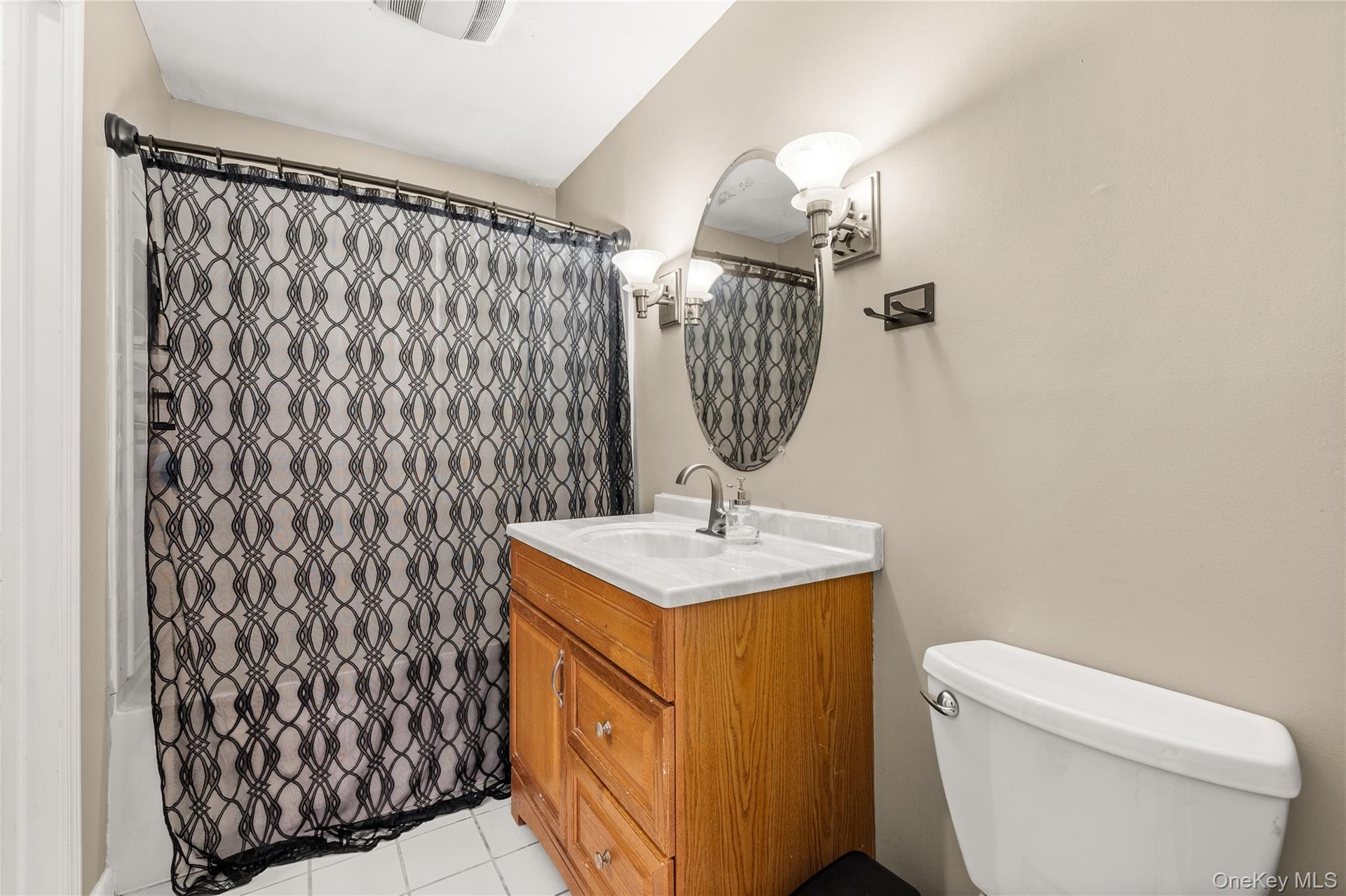 Oak finish vanity with a white countertop, oval mirror, wall-mounted light fixture, and white floor tiling
