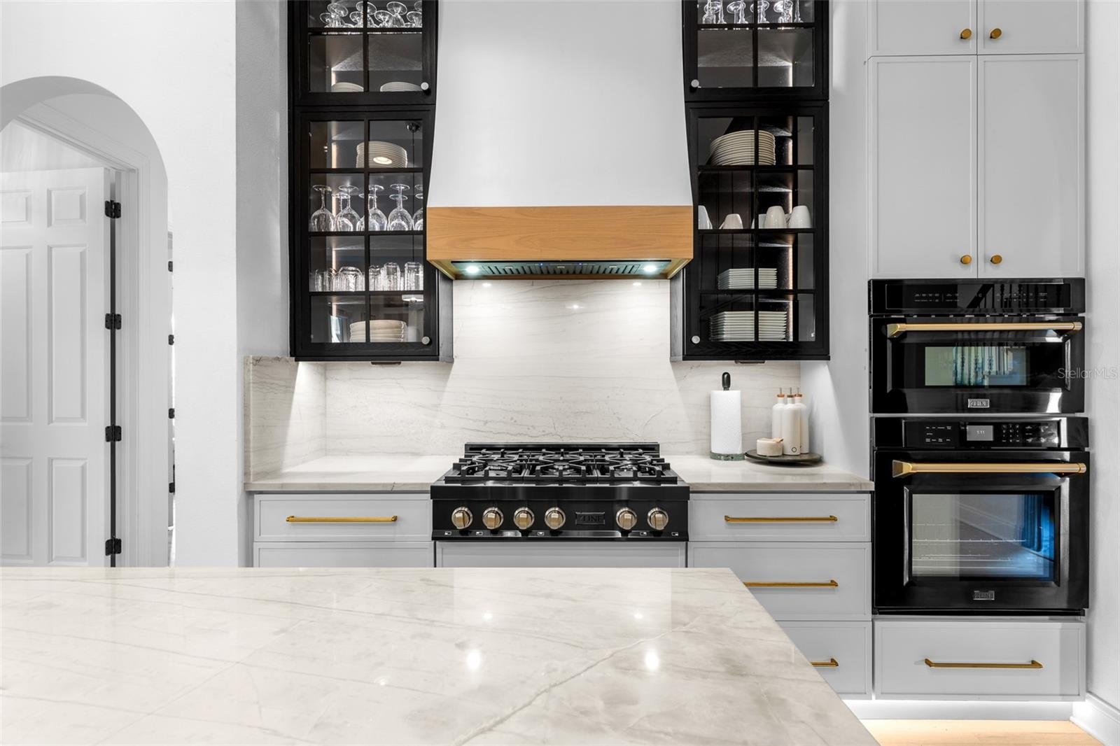 Gas range with wood-trimmed hood, glass-front black upper cabinets, and built-in wall oven and microwave.