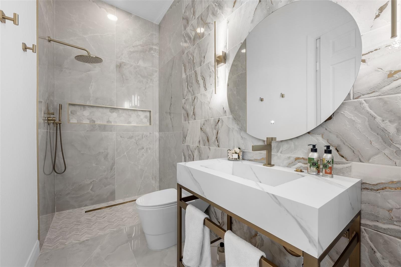 3rd Bathroom - Marble-look tile walls, console sink with brass frame, round mirror, and walk-in shower with brass fixtures.