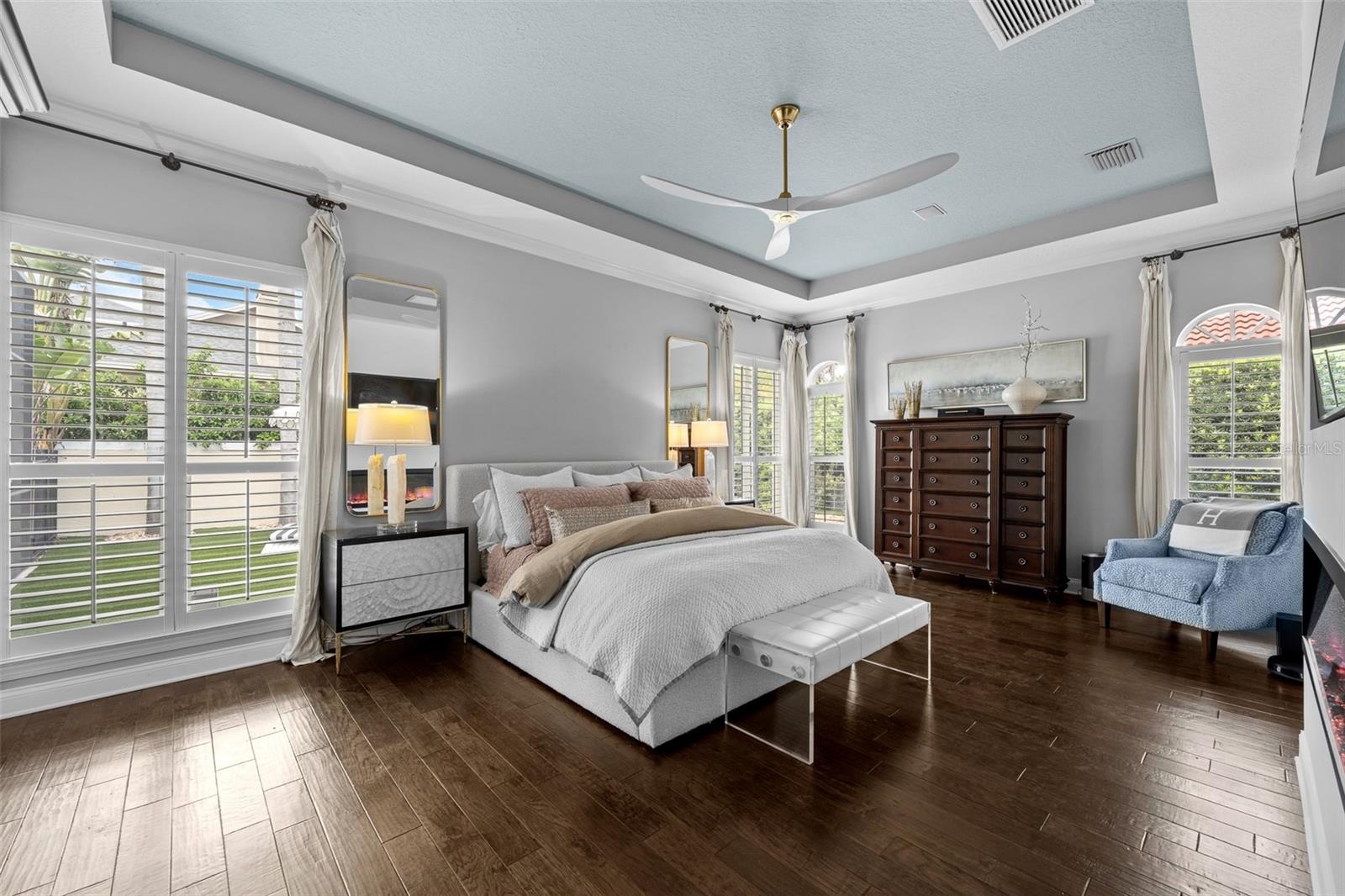 Tray ceiling with ceiling fan, dark wood flooring, and plantation shutters on multiple windows.