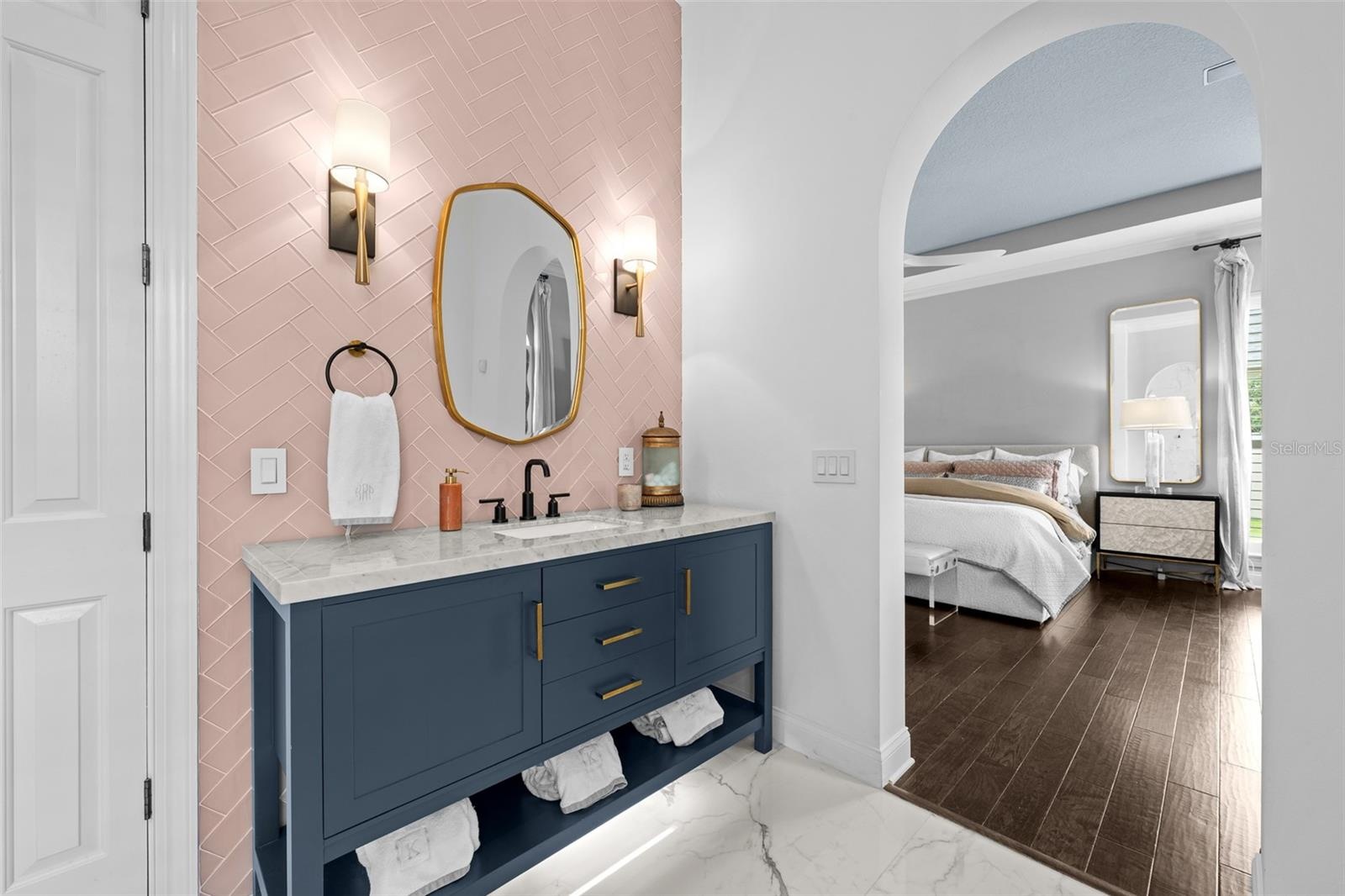 Navy vanity with stone top, pink herringbone tile wall, brass mirror, and arched view to the bedroom.