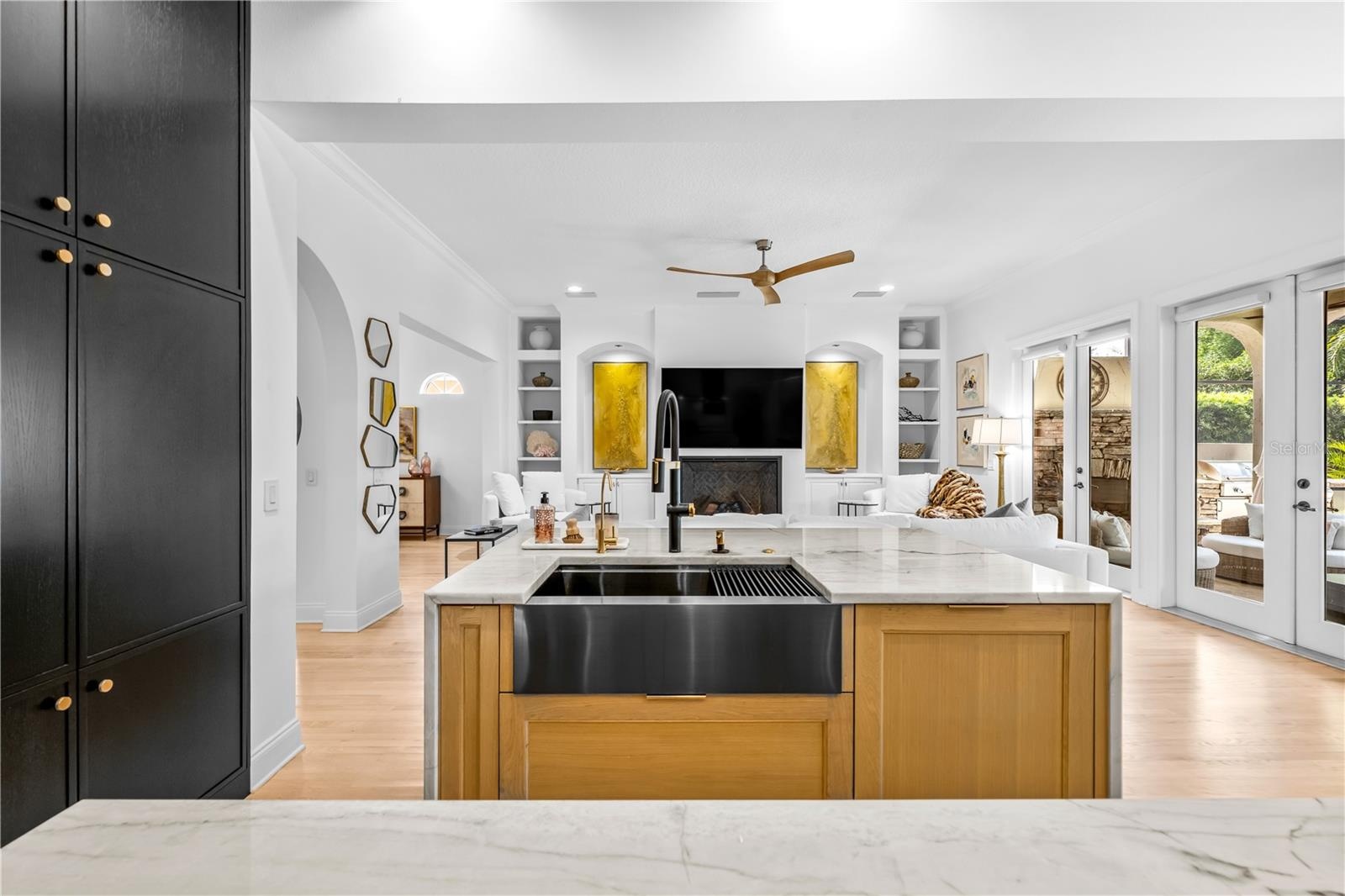 Farmhouse apron sink in a wood-front island, black tall cabinetry, and open sightline to the family room.