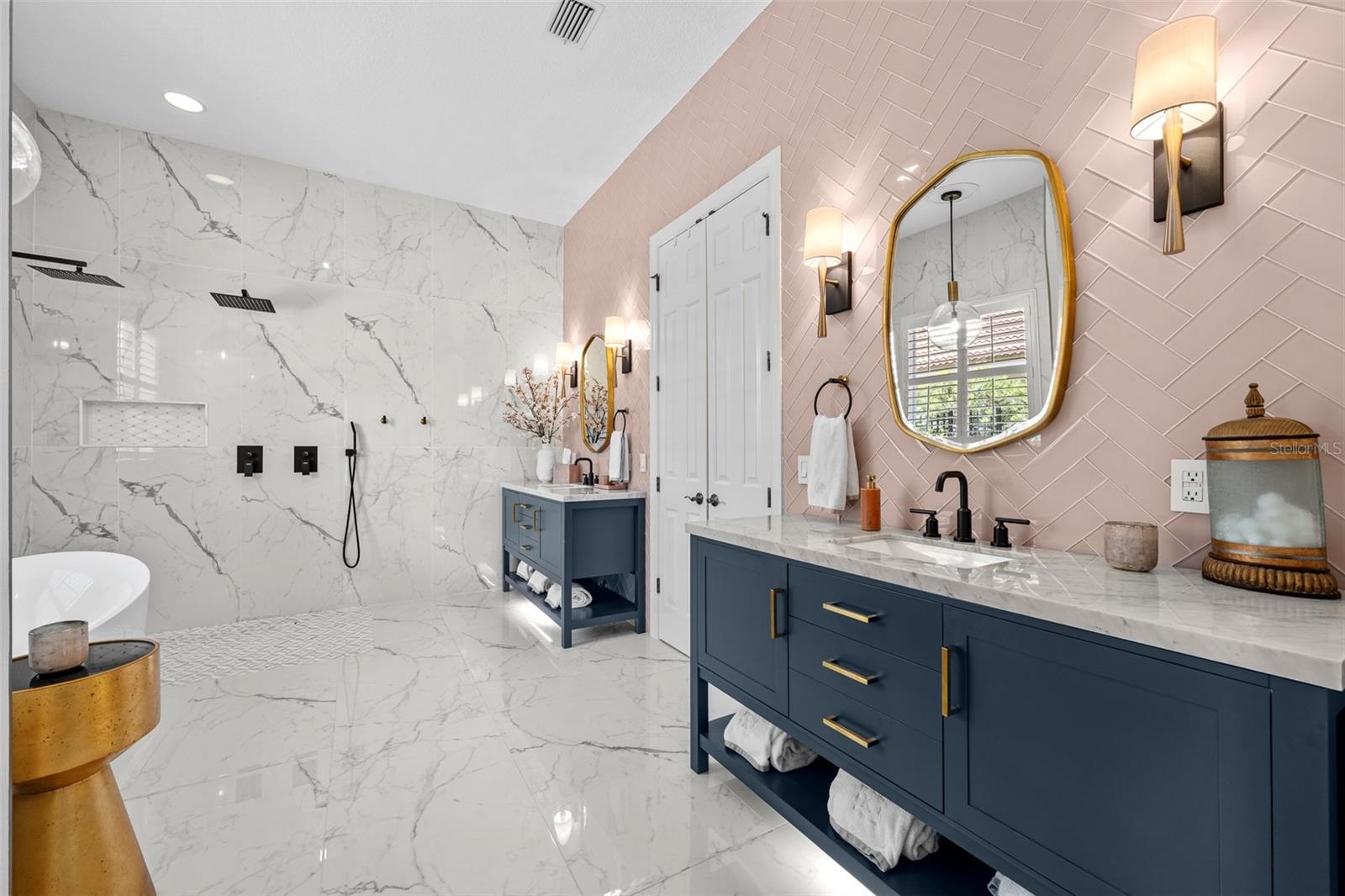 Primary Bathroom - Large-format marble-look tile, navy double vanity with brass hardware, and pink herringbone tile accent wall.