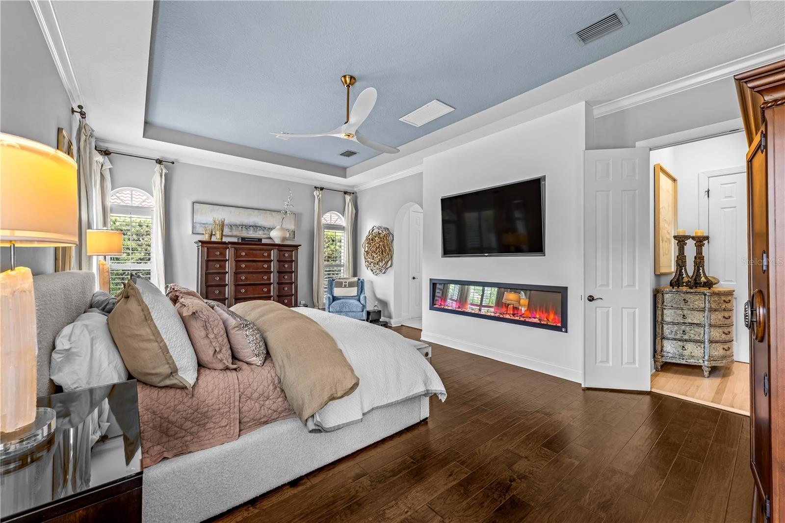 Tray ceiling, dark wood flooring, linear electric fireplace with mounted TV, and arched doorway.